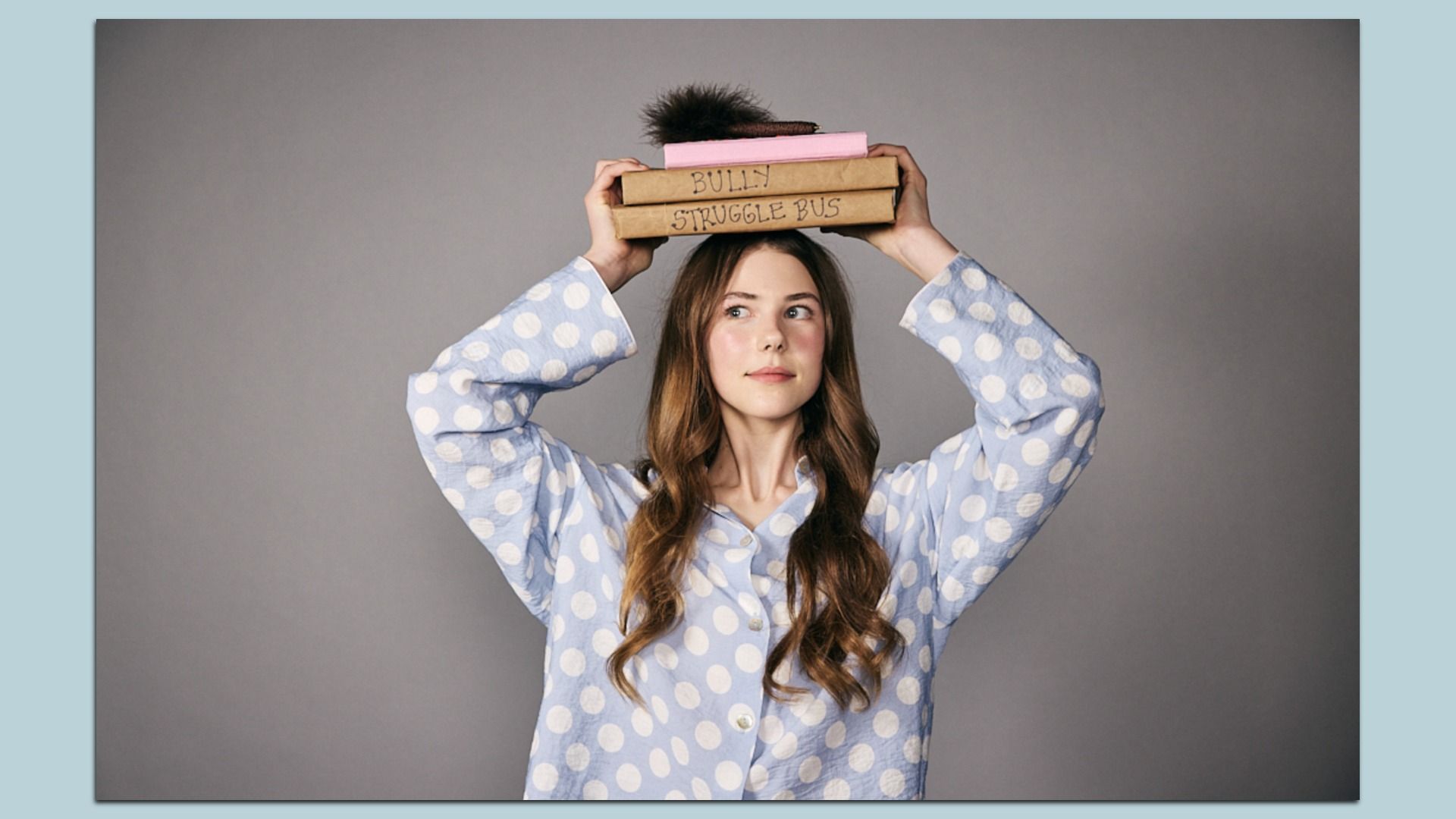 Young woman in a light blue polka-dot shirt balances a stack of books on her head; brown boxes labeled "BULLY" and "STRUGGLE BUS" against a gray backdrop.