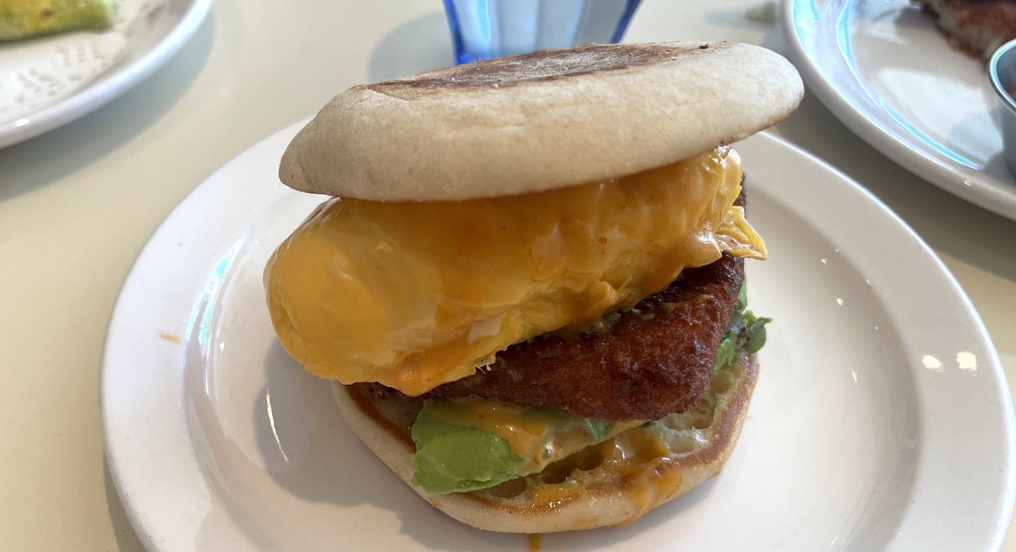 Close-up of a sandwich on a white plate: a toasted bun over a thick, crispy fried chicken patty with melted cheddar cheese, lettuce, and orange sauce dripping from the sides.