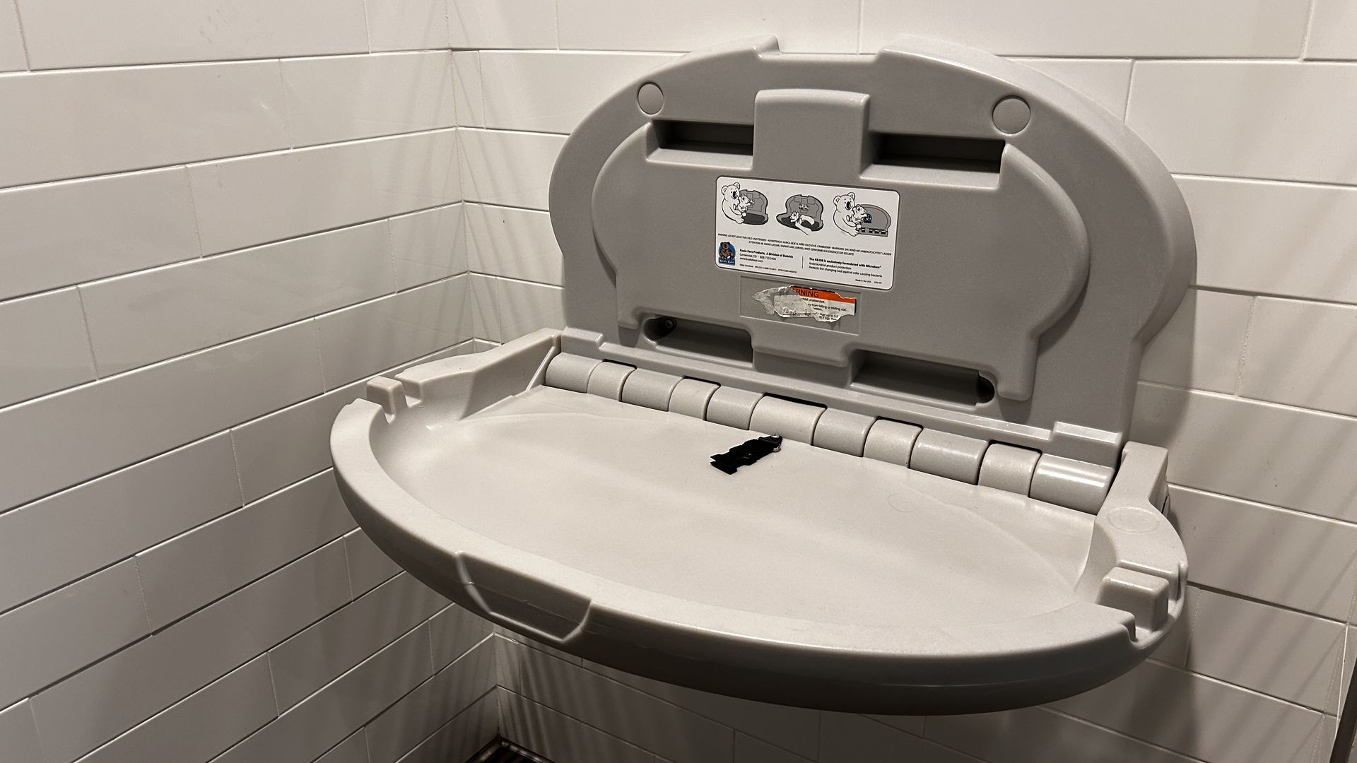 A gray plastic changing table is open in a bathroom with white-tiled walls.