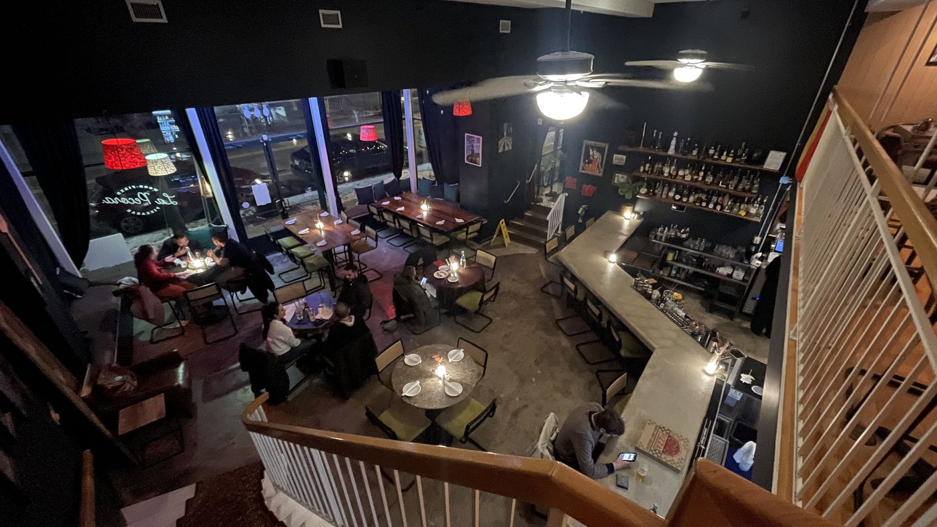 Cozy dimly lit bar with wooden tables and chairs, a long bar counter, patrons seated at tables and bar, large windows, ceiling fans, and bottles displayed on wall shelves.