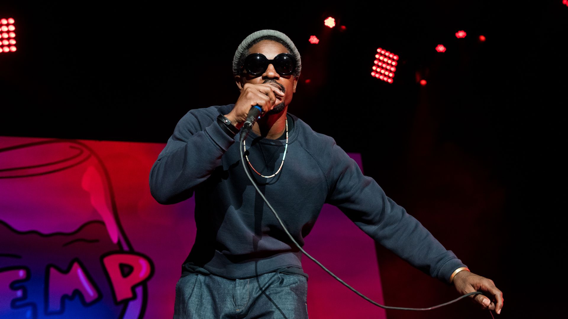 A man wearing dark clothes and large wraparound sunglasses raps on stage