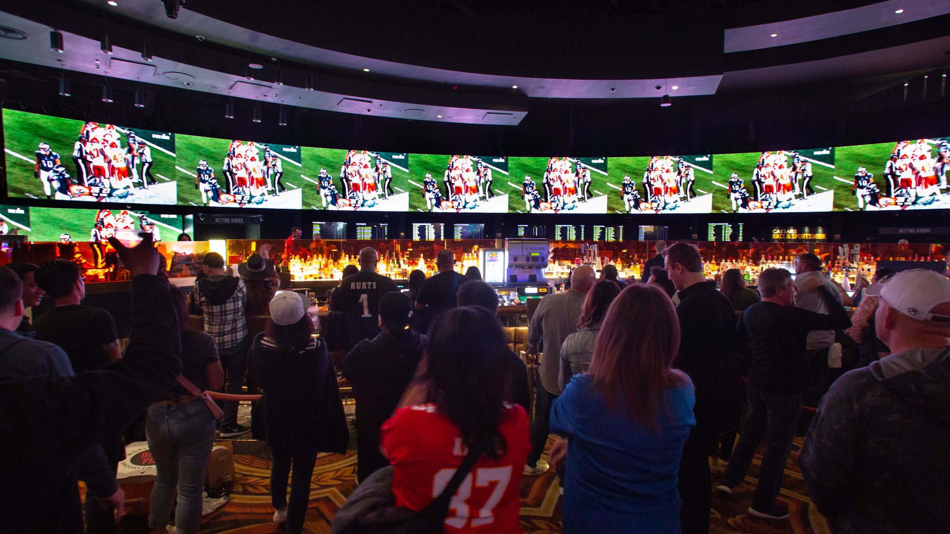Crowd watching football game on multiple screens above a bar with bright orange lighting, some wearing jerseys like a red #87 and black Hurts #1, in a dimly lit sports bar.