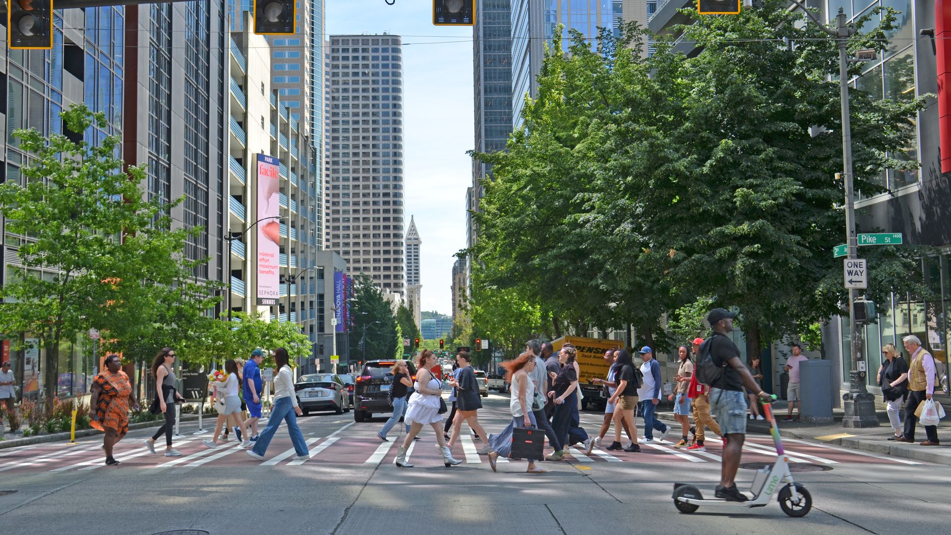 Busy city crosswalk with pedestrians walking across street among tall buildings, green trees, and traffic lights on a sunny day at Pike and 2nd. 