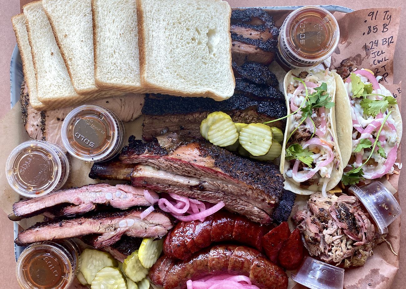 Barbecue platter with sliced brisket, sausages, pulled pork, two tacos with cilantro and onions, sliced white bread, pickles, pickled red onions, and several small containers of barbecue sauce on brown paper.
