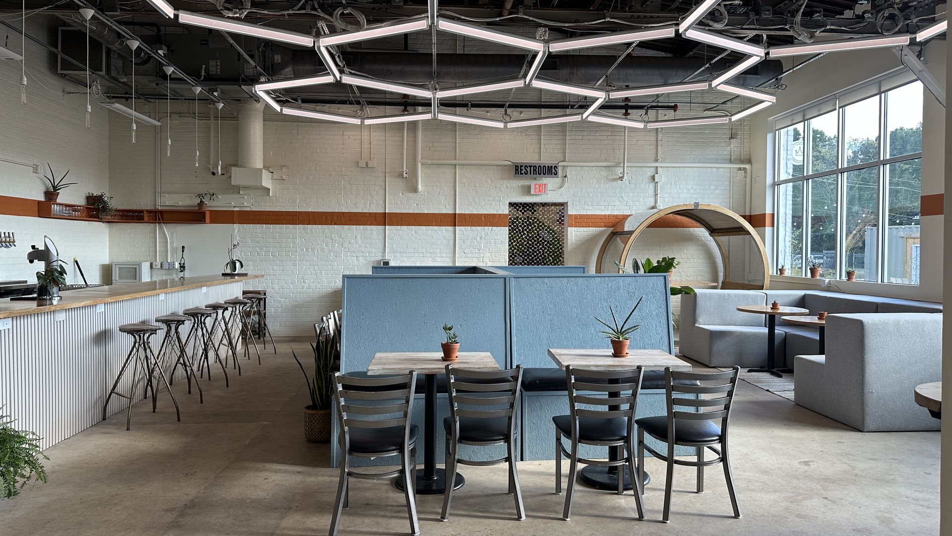 Industrial cafe interior with a white brick wall, honeycomb-shaped LED light fixtures overhead, a long bar with stools on the left, blue dividers, potted plants, and large windows to the right. And/or cafe is a new concept from former Lorem Ipsum owner that opens in Belmont neighborhood in the former Change Please cafe space inside Innovation Barn.