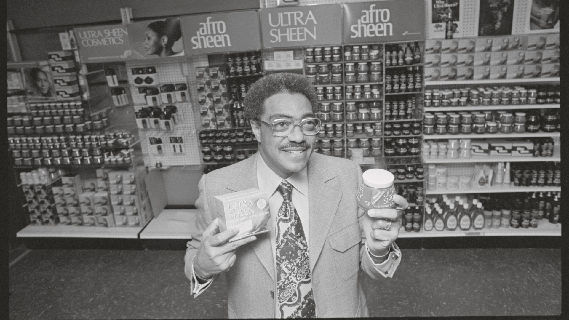 Black-and-white image of a smiling man in a suit in a store aisle, shelves filled with Afro Sheen and Ultra Sheen hair products; he holds a boxed product and a can.