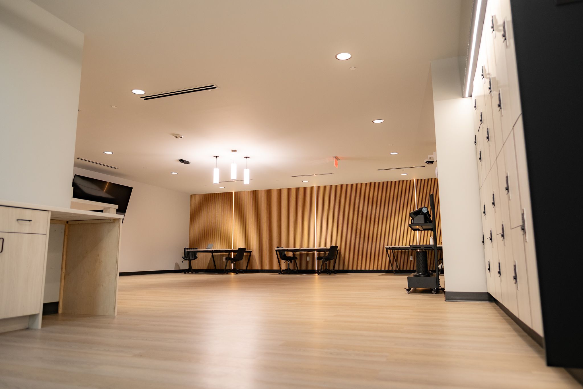 Empty modern interior with light wood floor, white walls, and ceiling lights. A vertical wooden slat wall at the far end, with tables and chairs, and right-side lockers.