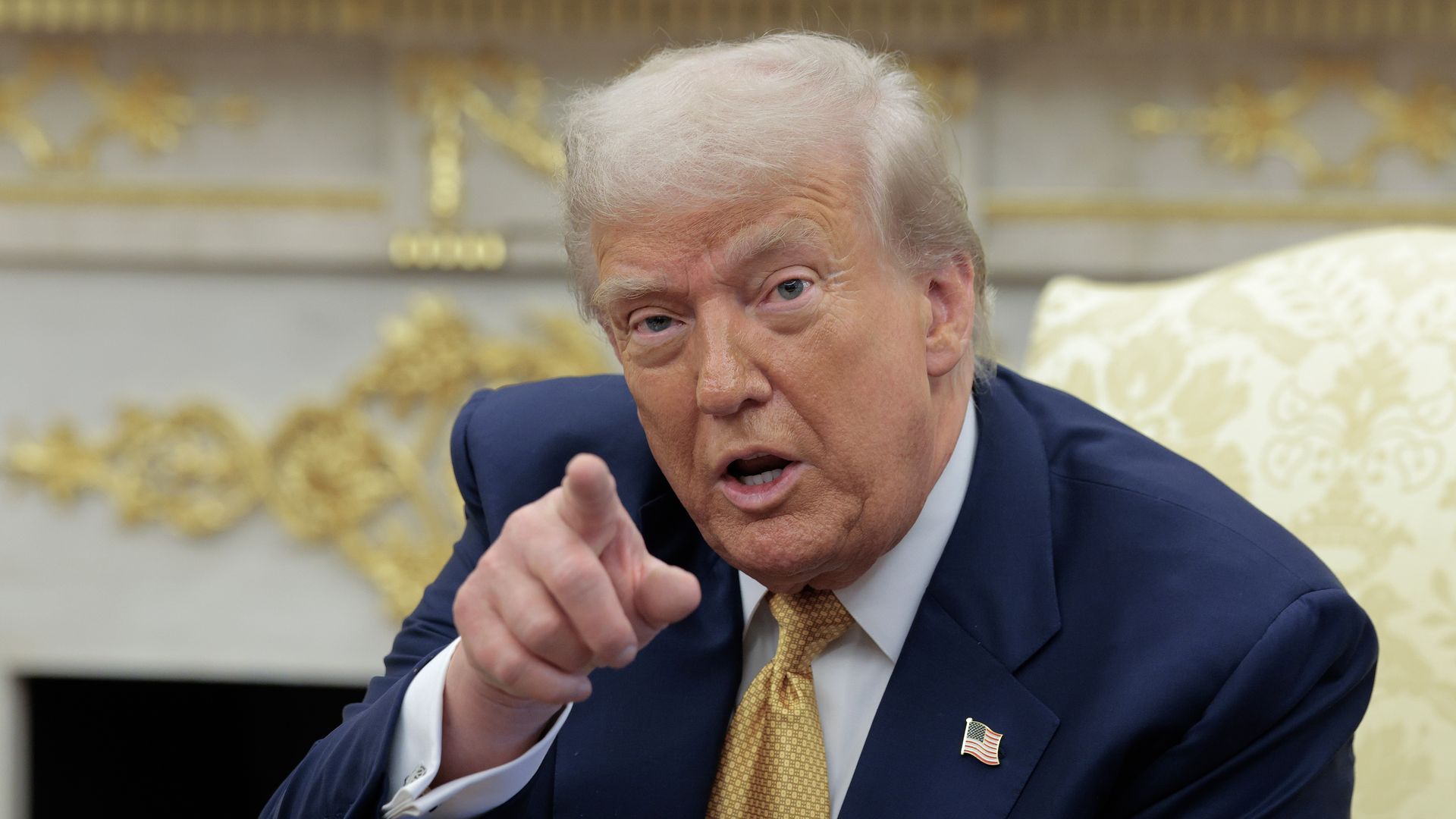 President Trump, with light hair wearing a navy suit, gold tie, and American flag pin points forward with a serious expression against a background with gold decor.