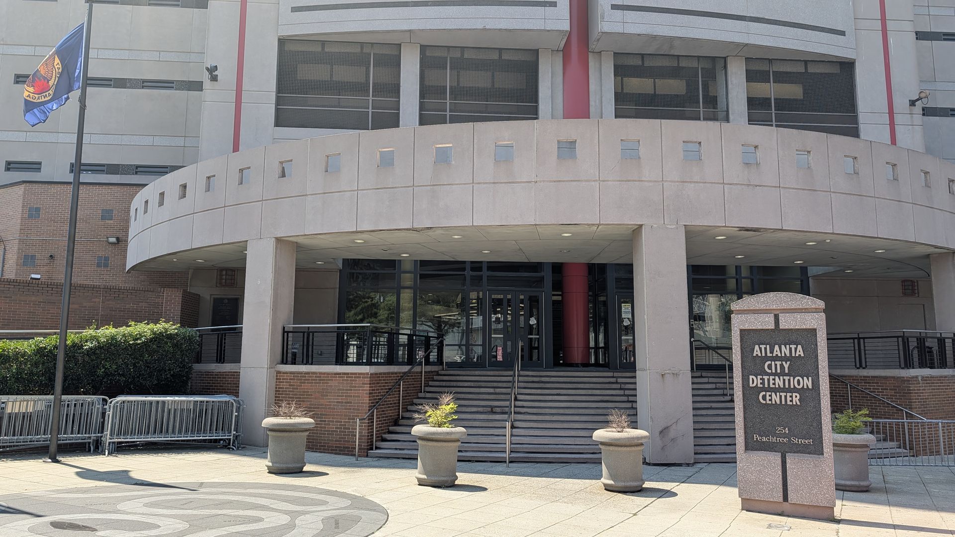 Exterior of the Atlanta City Detention Center: curved concrete facade with small square windows, red columns, glass doors, a flagpole with a blue flag, and a granite sign reading "Atlanta City Detention Center".