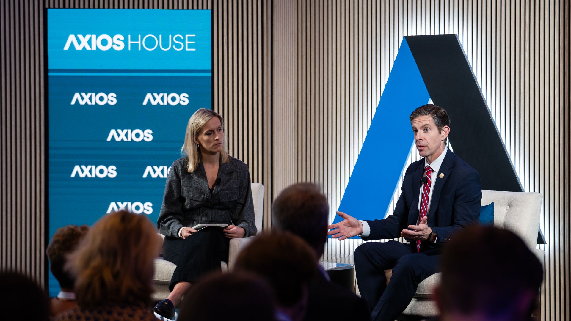 Discussion on a stage with Axios branding. A woman in a gray blazer sits on the left with a notebook; a man in a dark suit and red striped tie speaks on the right. A blue Axios screen is behind them.