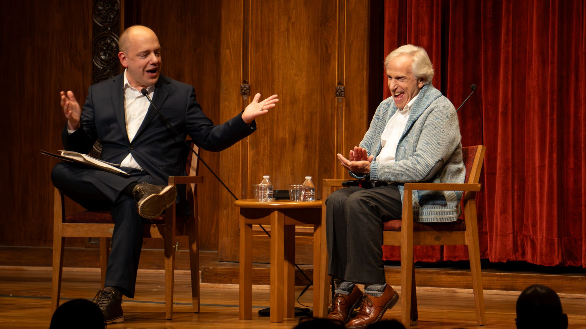Two men engaged in conversation on a stage with wooden walls and red curtains; one in a black suit speaking, the other older in a light blue sweater laughing; small table with water bottles between them.