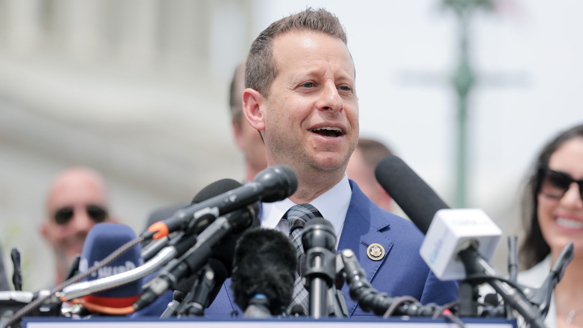 A man in a blue suit and tie speaks at a podium during an outdoor press conference, surrounded by many microphones.