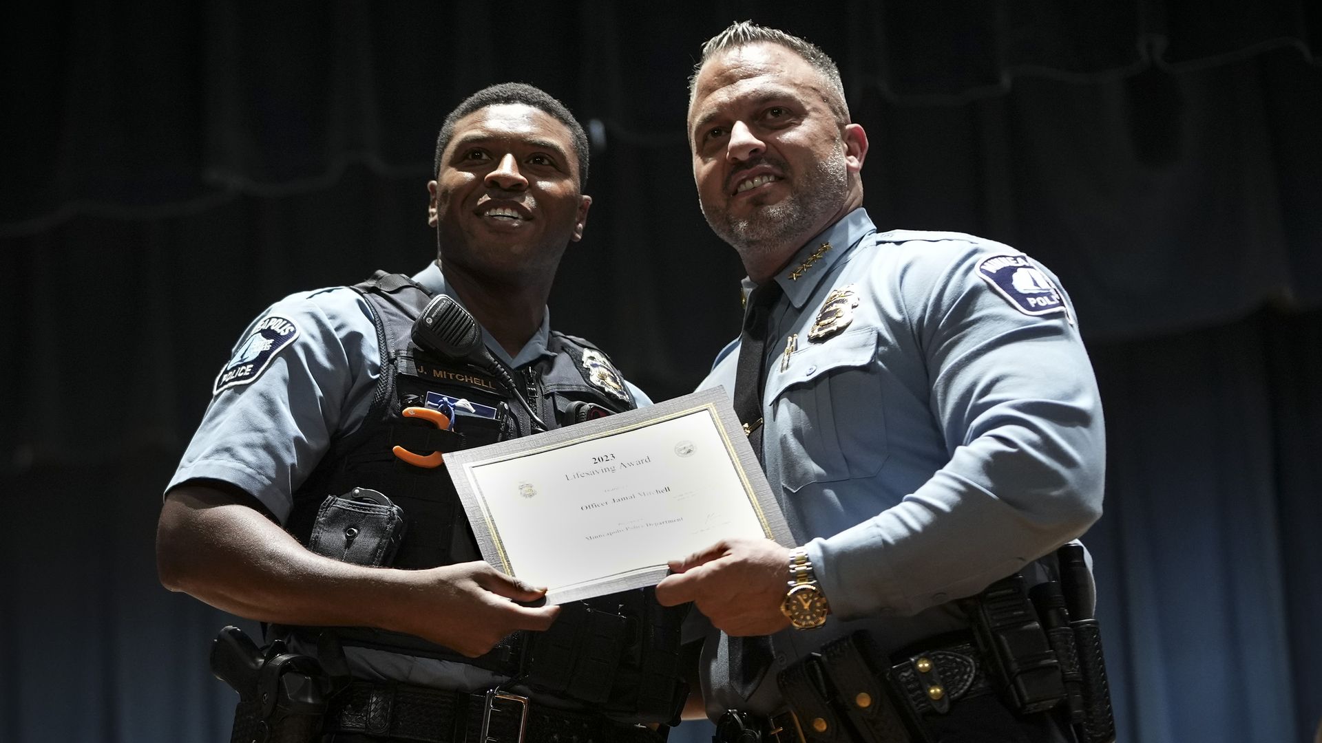 Two police officers on a stage exchange a framed certificate; both wear light-blue uniforms with badges and gear. A dark curtain backdrop and a seated woman to the right.