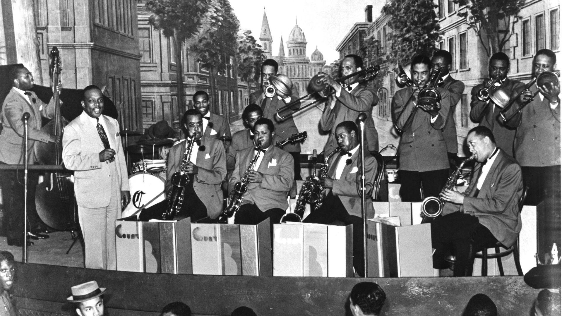 Black-and-white street scene of a suit-wearing brass band performing; saxophones and trumpets players, crowd in foreground, urban buildings and trees lining the avenue.