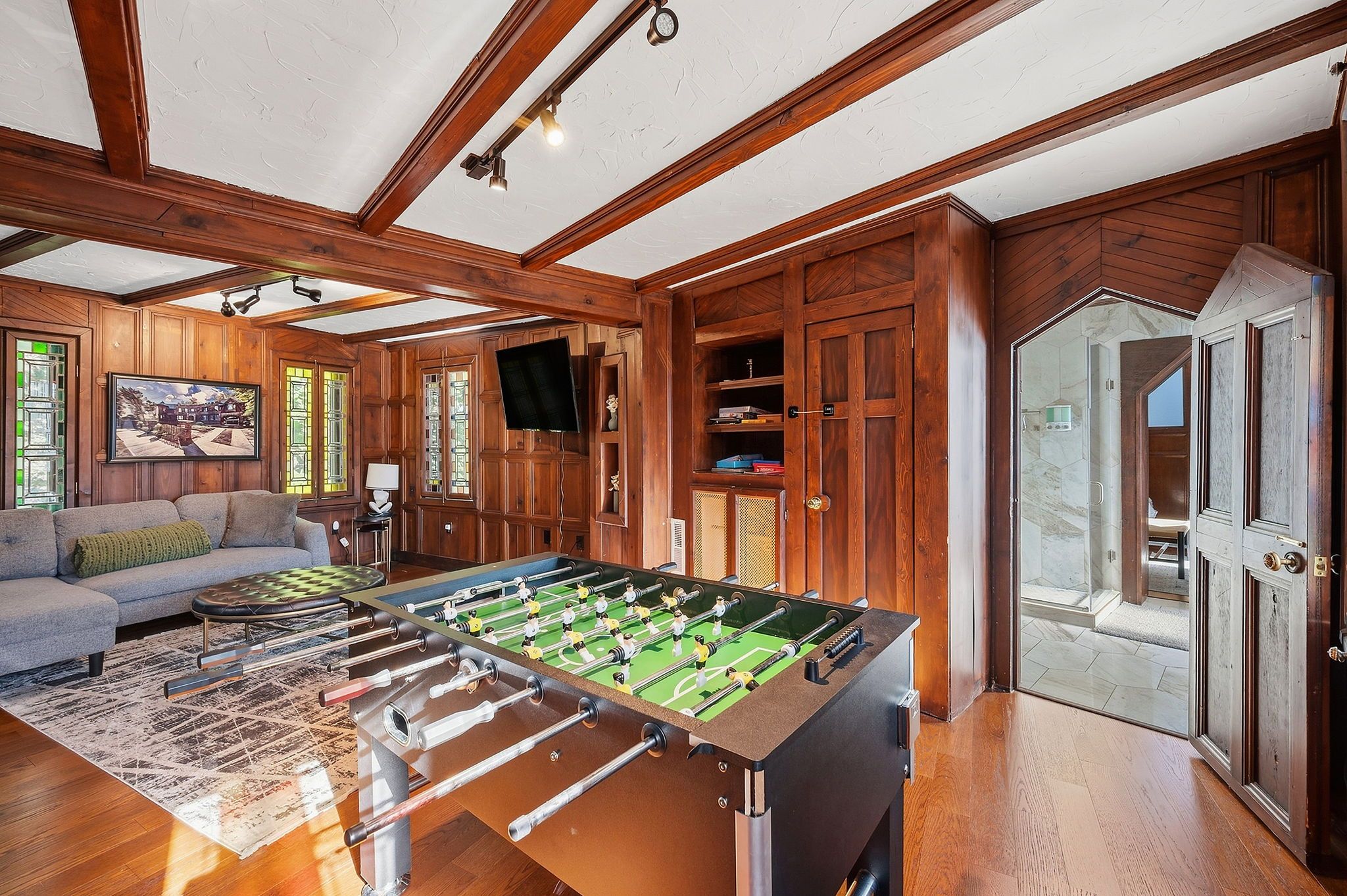 Wood-paneled living room with exposed ceiling beams, a foosball table and a gray sectional sofa. Built-in shelves, a wall TV, stained-glass windows, and a marble-tiled bathroom doorway.