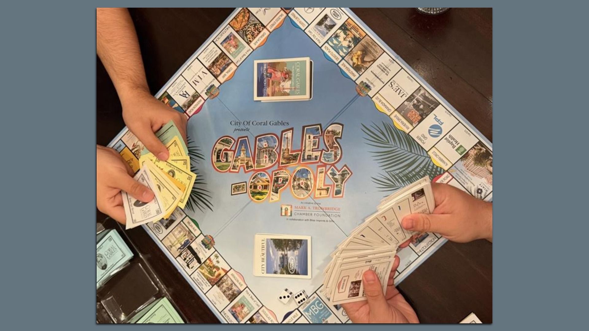Two people playing a custom board game called Gables-opoly by the City of Coral Gables, with colorful cards and game money on a wooden table.