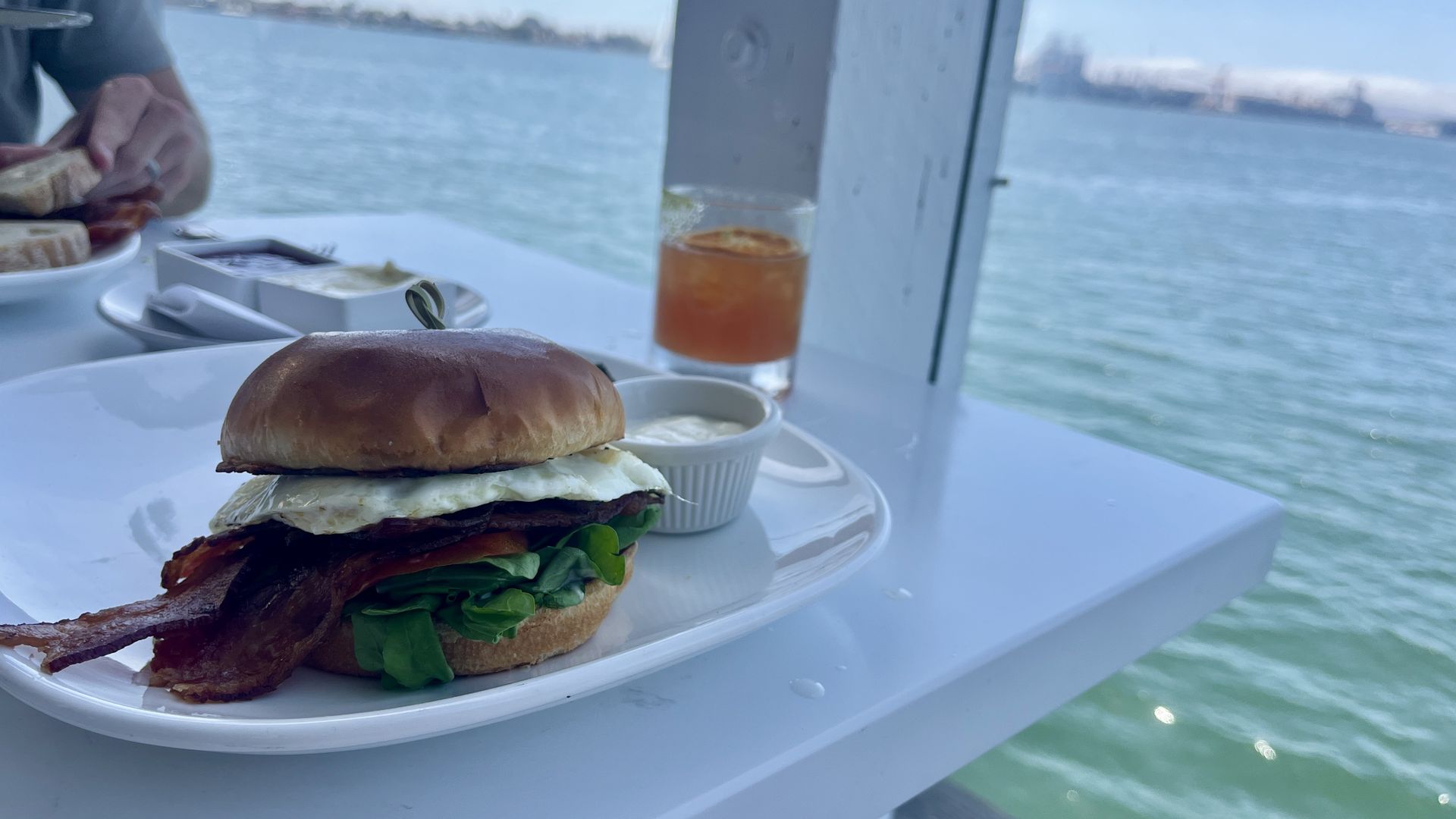 Bacon, egg, tomato and lettuce sandwich on a white plate with dipping sauce, near a window overlooking blue water under a clear sky.