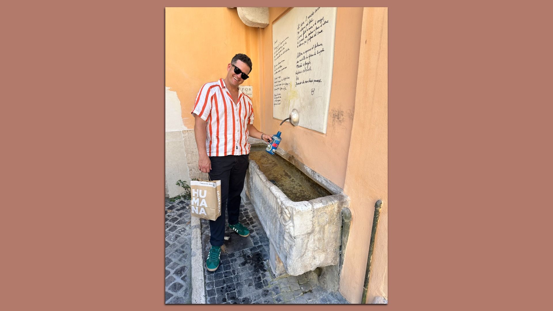 Man in a red-and-white striped shirt and sunglasses stands beside a stone water trough on an orange wall, holding a blue bottle and a beige HUMANA bag.