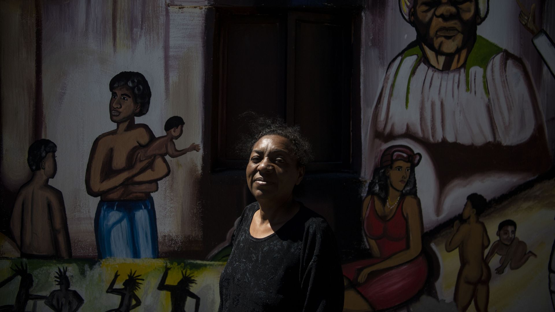 A woman wearing black stands in front of a large mural, half of her face obscured by shadows. 