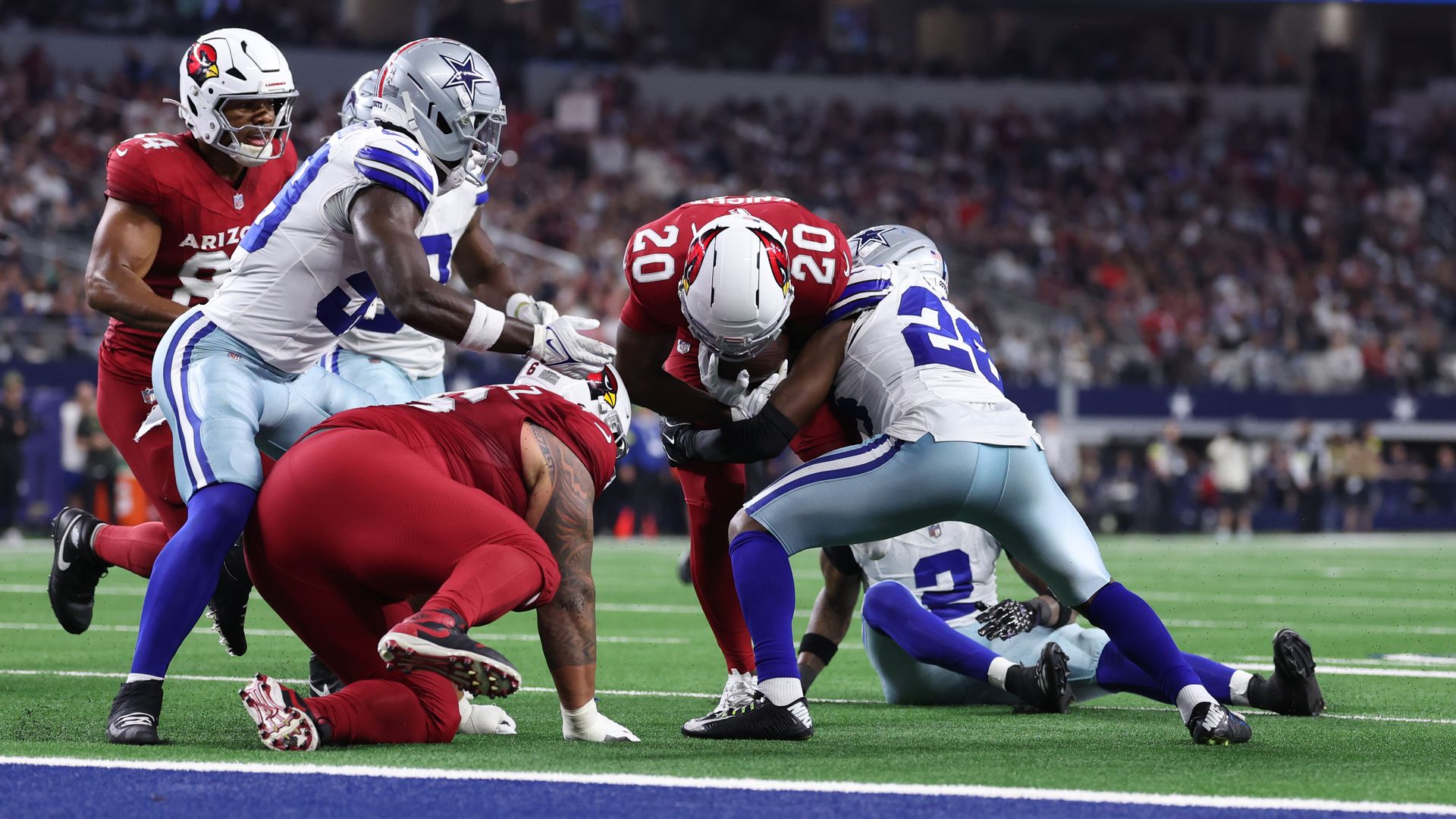 Arizona Cardinals player in red jersey being tackled near the end zone by multiple Dallas Cowboys players in white and blue uniforms during a football game.