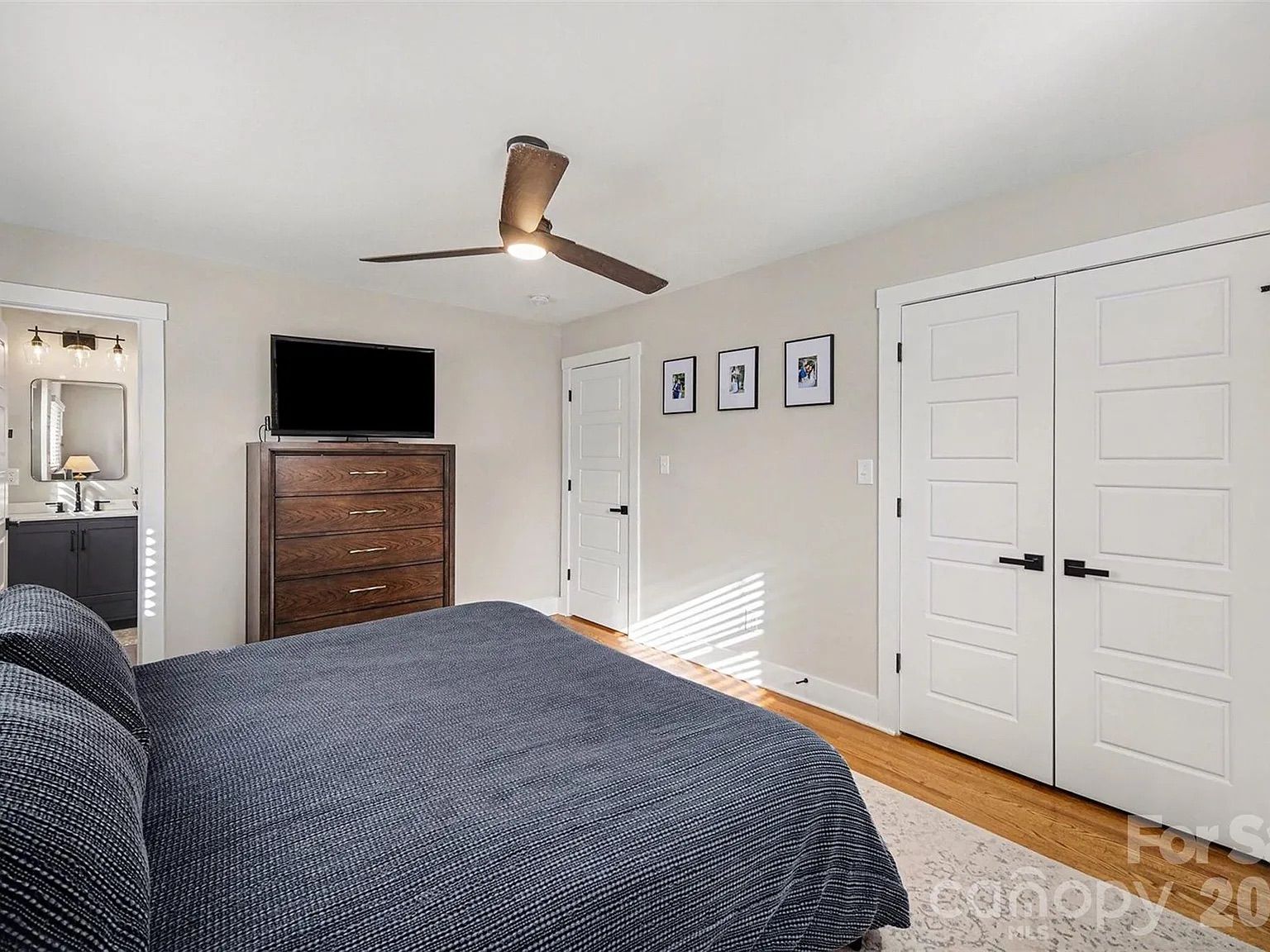 Bedroom with gray bedspread, wooden dresser with TV, white walls, ceiling fan, three framed photos, two white doors, and hardwood floor with rug.