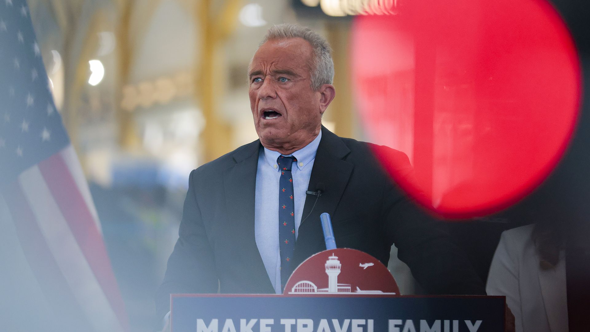 Man with gray hair and blue eyes in dark suit speaks at podium with sign reading "MAKE TRAVEL FAMILY FRIENDLY AGAIN," American flag on left, large red circle to right.