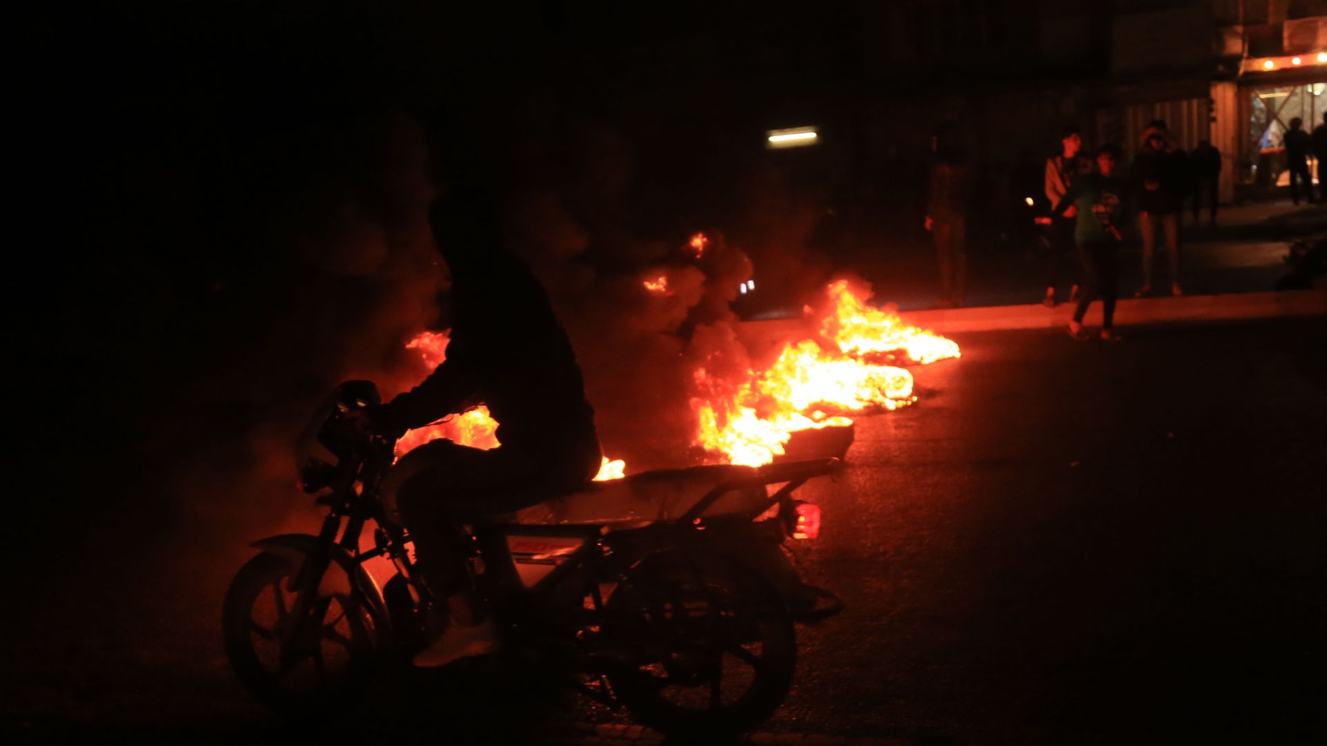 A person riding a bike with a protest in the dark and fire in the background.