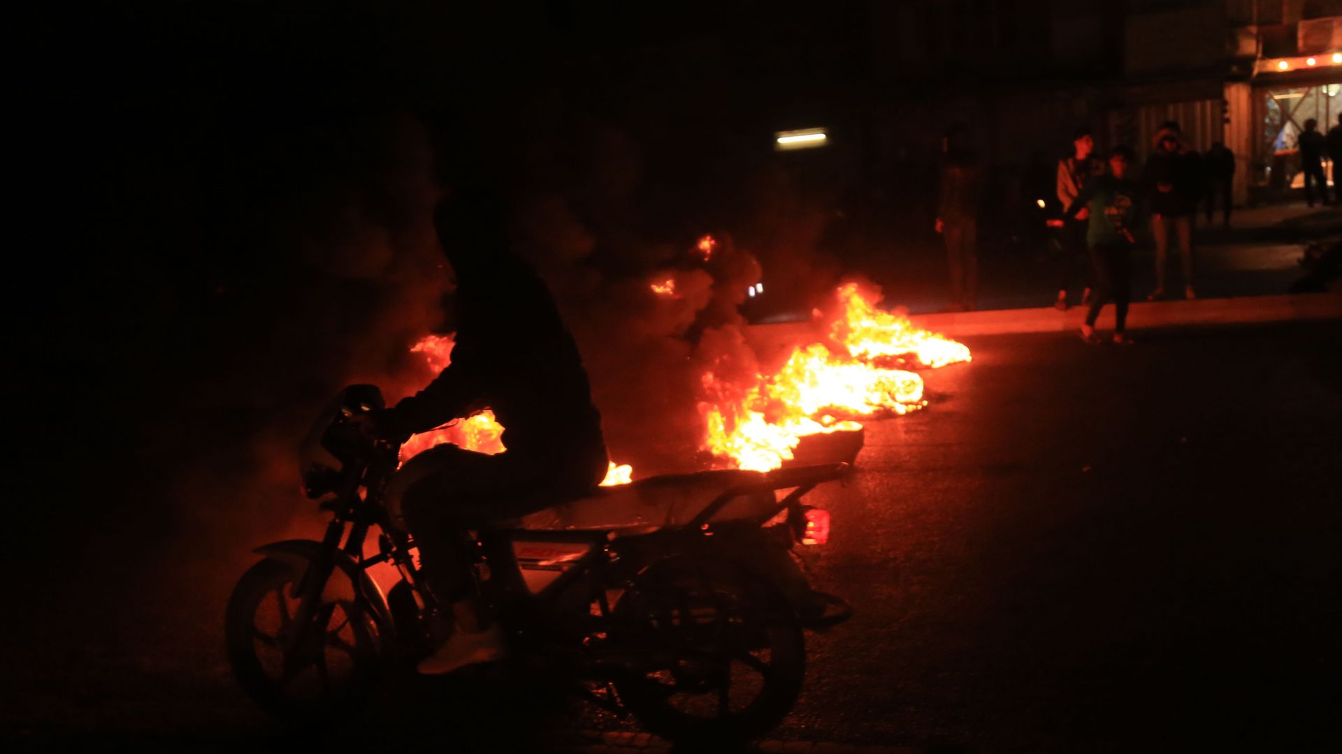 A person riding a bike with a protest in the dark and fire in the background.