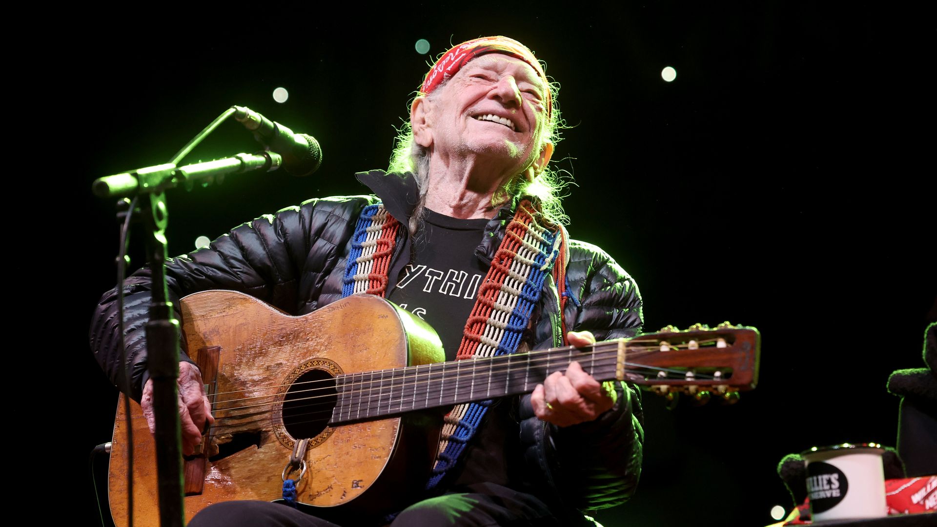 Willie Nelson smiles and sits while playing an acoustic guitar in a darkened music venue