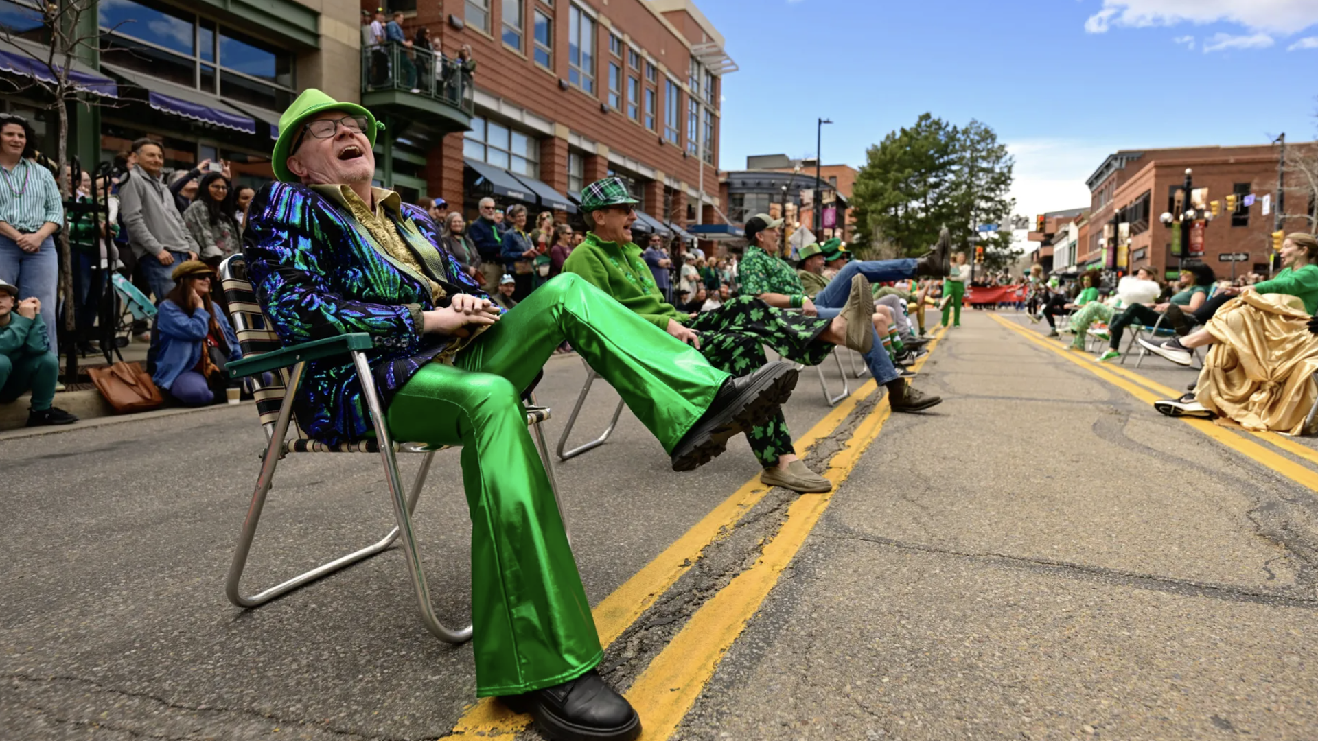 A lively street parade with spectators lining a city block. In the foreground, a man in a bright green suit laughs while seated in a folding chair, others in green outfits kick their legs and cheer.