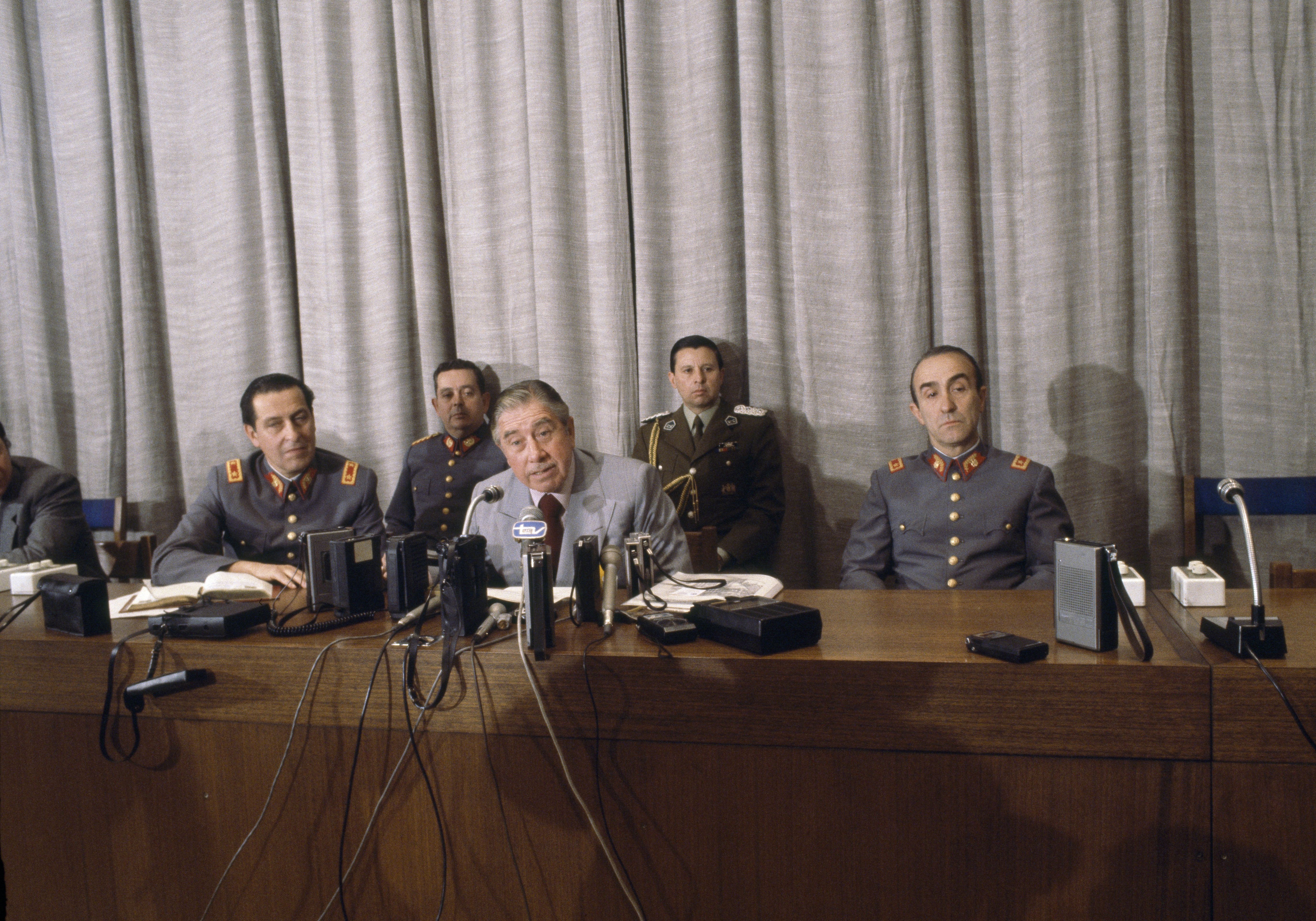 Chilean dictator Augusto Pinochet sits at a long wooden desk with several microphones in front of him as he speaks during an news conference