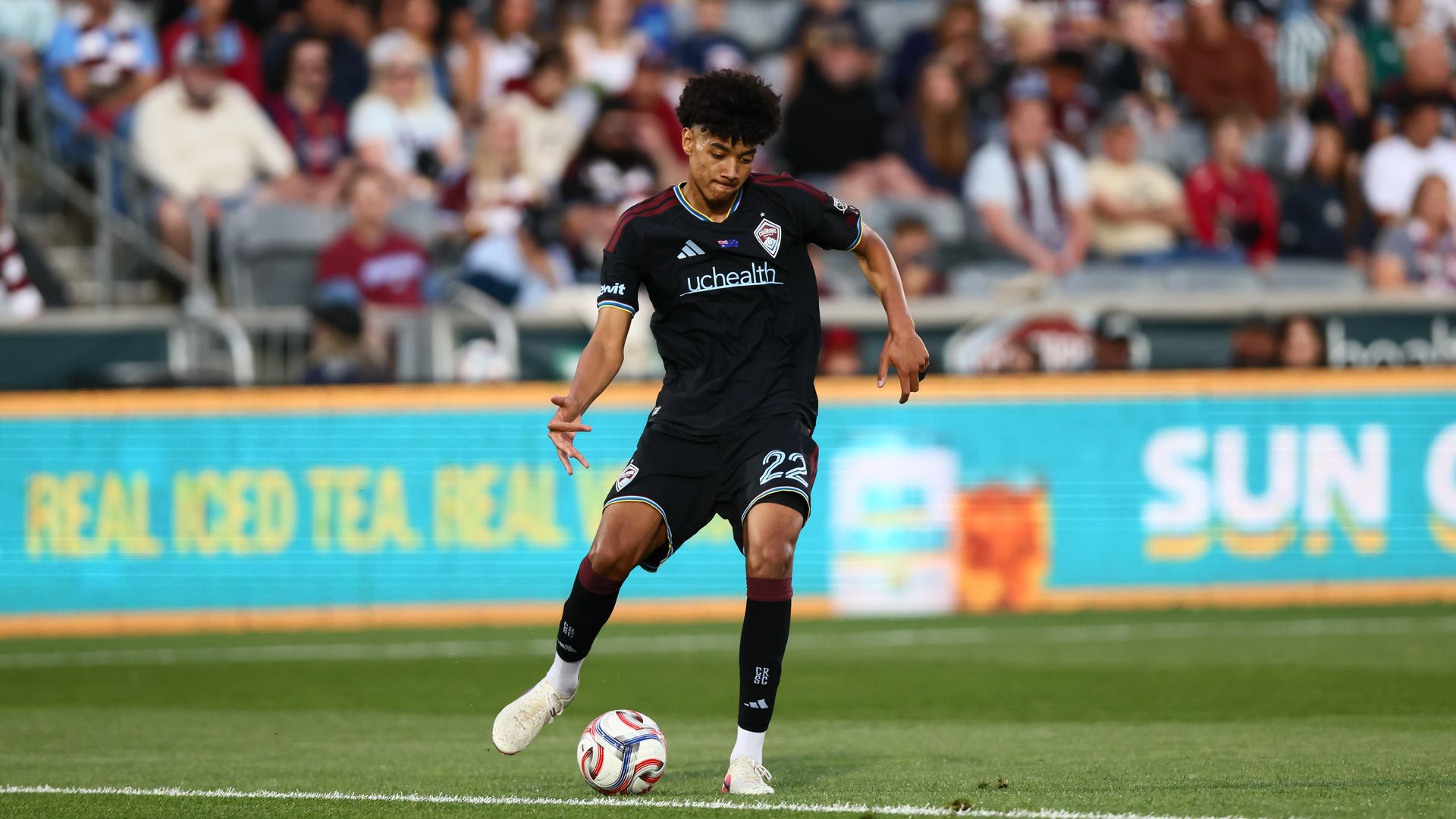 Soccer player in a black kit with blue trim and uchealth sponsor dribbles a ball on a green pitch, with a large, blurred crowd in the stands behind.