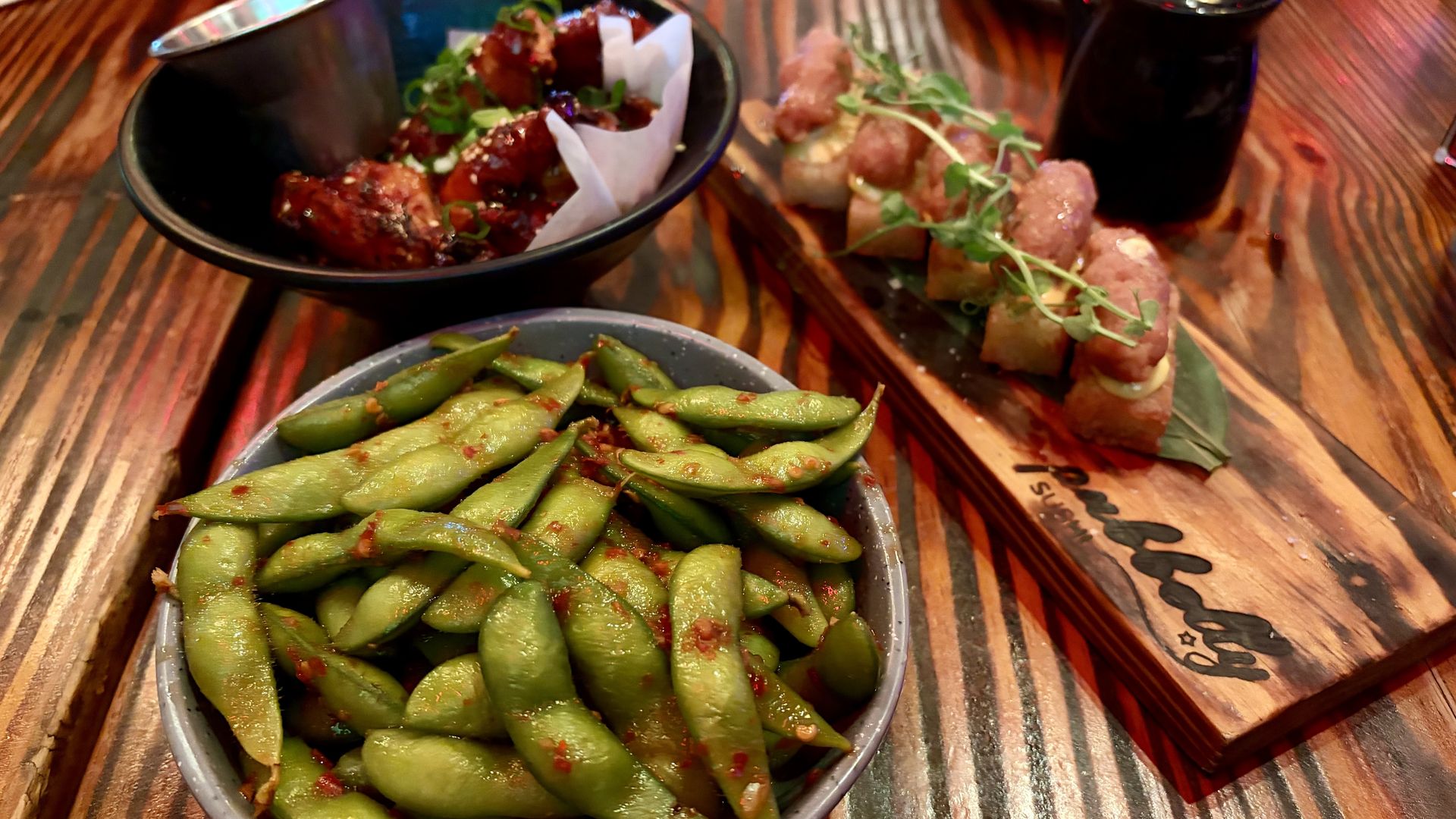 Three dishes on a wooden table: a bowl of spicy edamame, a bowl of glazed chicken wings garnished with green onions, and a wooden board with sushi topped with sauce and microgreens.