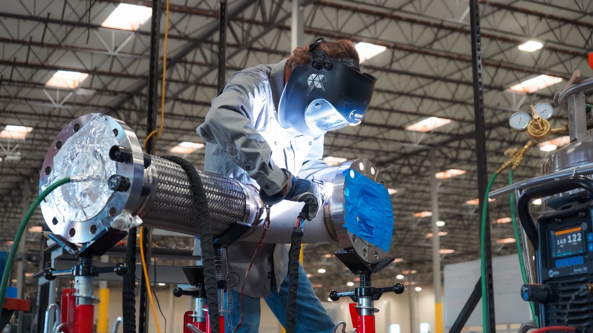 A welder with a dark helmet and gray work shirt welds a large metal pipe in an industrial workshop, with gauges, hoses, and exposed steel trusses in the background.