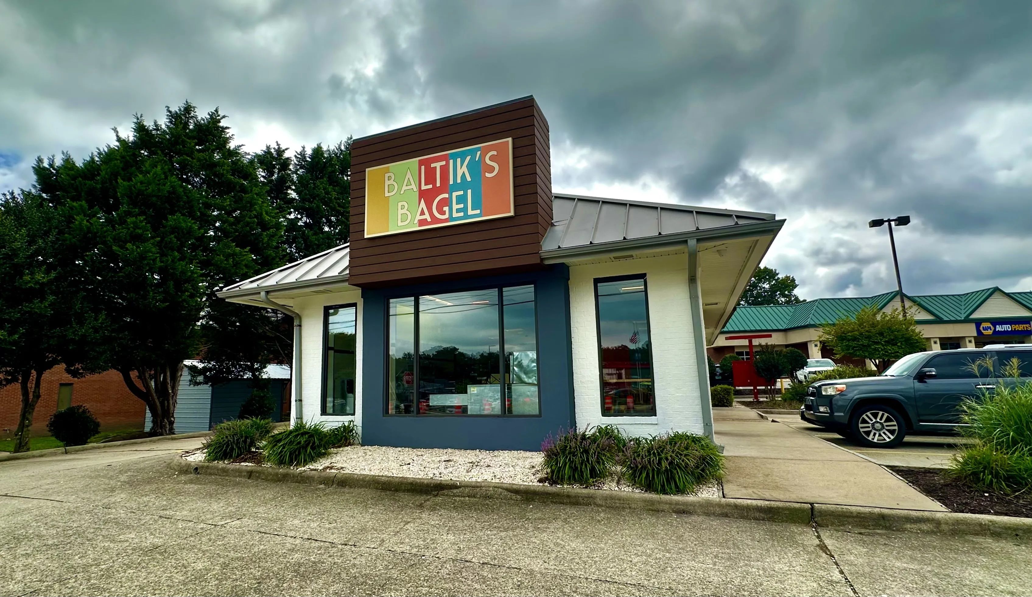 Colorful sign reading Baltik's Bagel atop a brown storefront with white brick and blue trim; large front windows, cloudy sky, trees to the left, and a blue SUV in the parking lot.