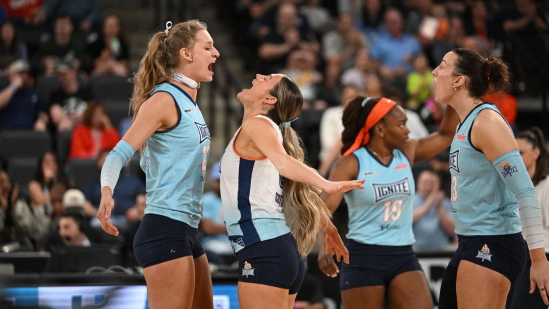 Four female volleyball players in light-blue uniforms celebrate on a court, with a cheering crowd in the background.