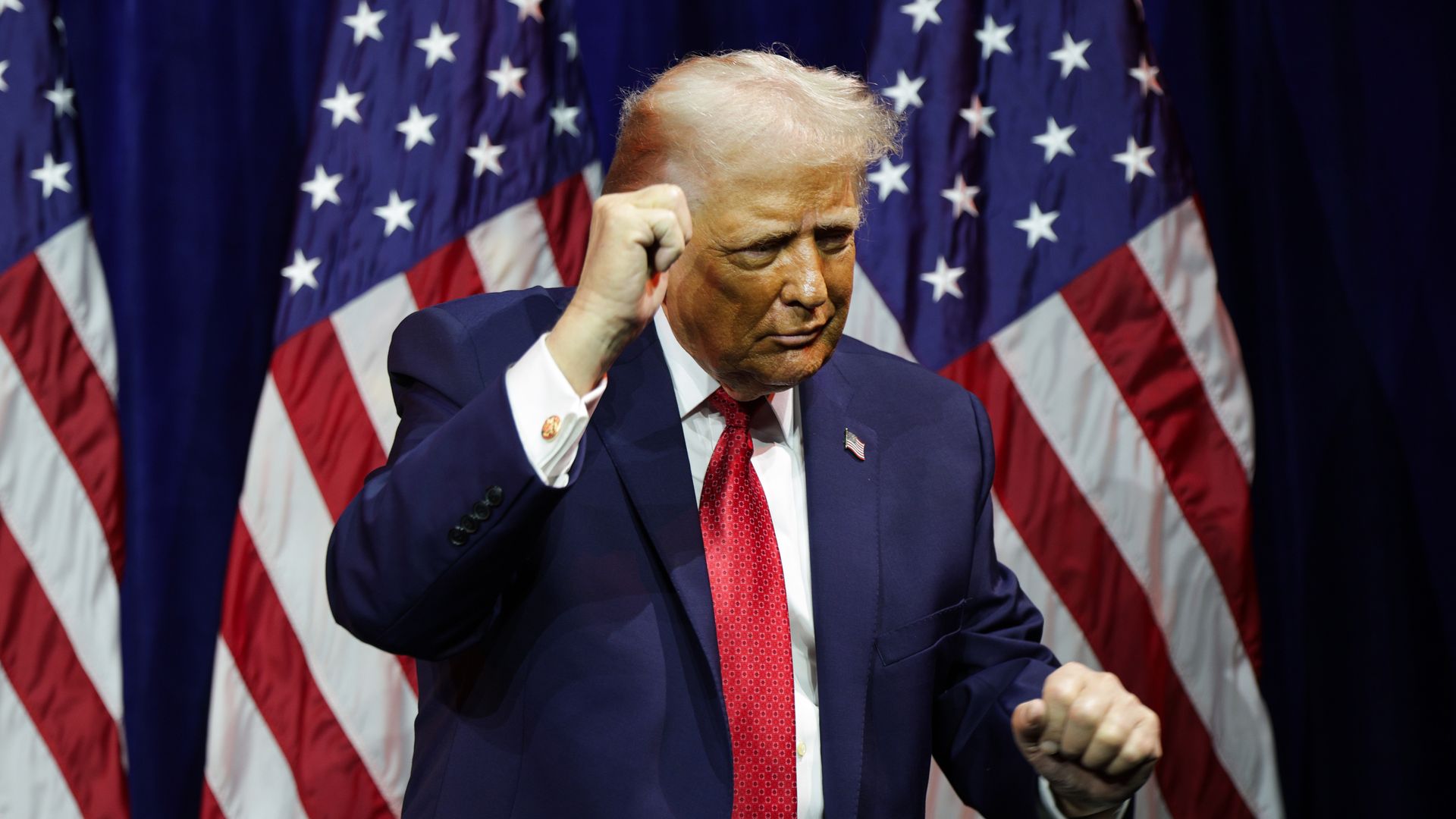 Trump standing in front of three American flags with his fist raised while wearing a navy suit jacket, white collared shirt and red tie