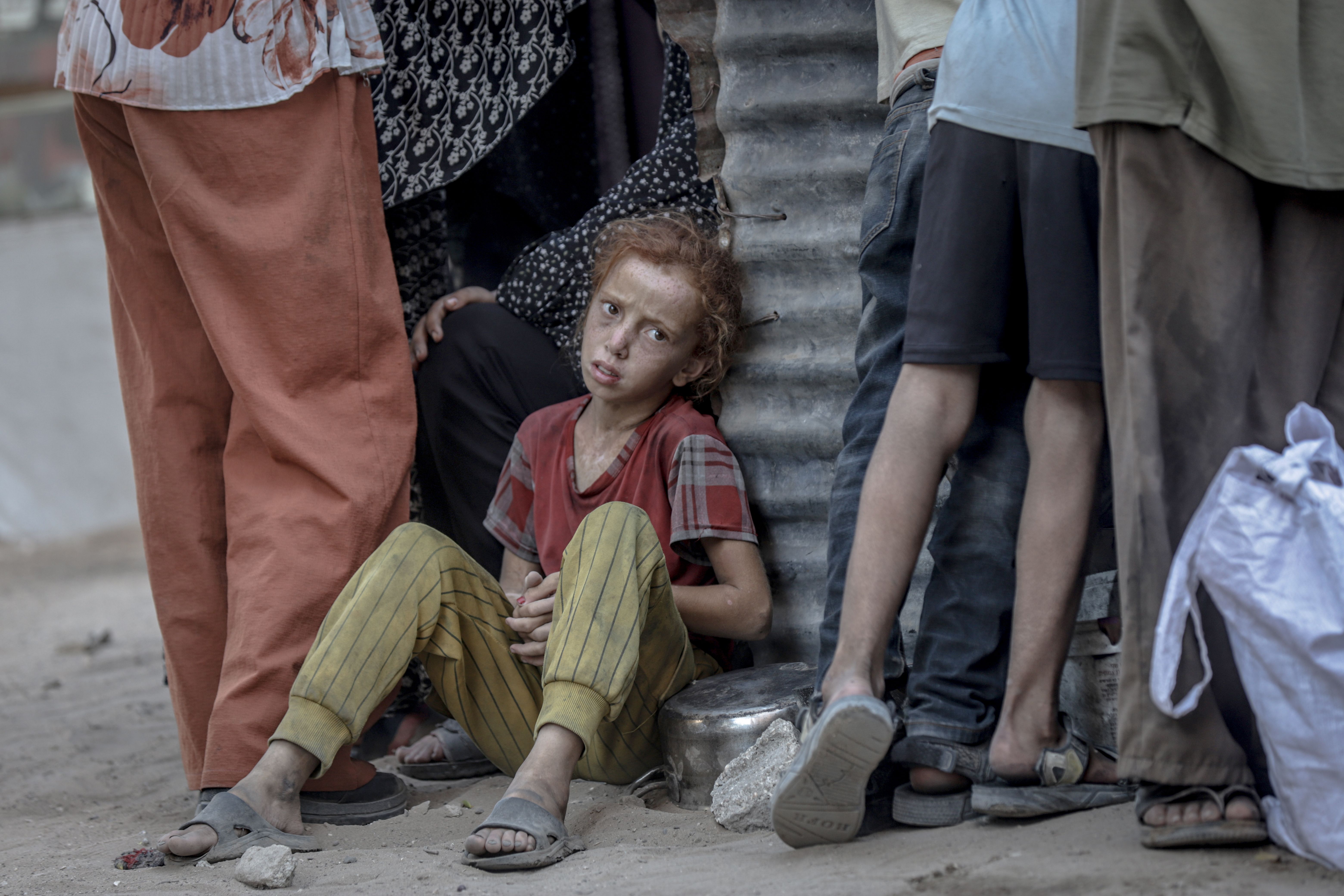 A young, red-haired child in a red shirt and yellow striped pants sits on the ground, leaning against a rusty metal sheet, surrounded by standing people wearing casual clothes and sandals.