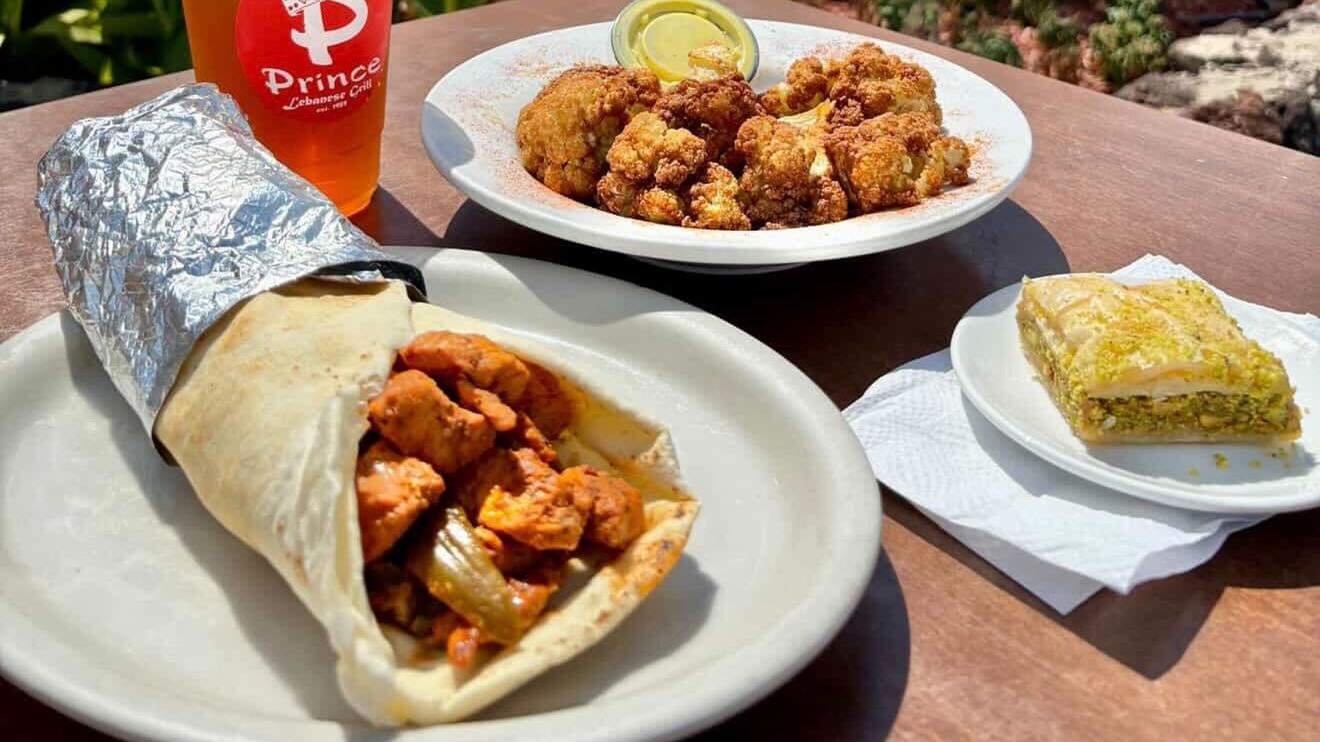 A close-up photo of a pita wrap filled with chicken shawarma on a plate, a plate with baklava and another plate in the background with fried cauliflower. A clear plastic cup of iced tea can be seen behind with a sticker saying Prince Lebanese Grill