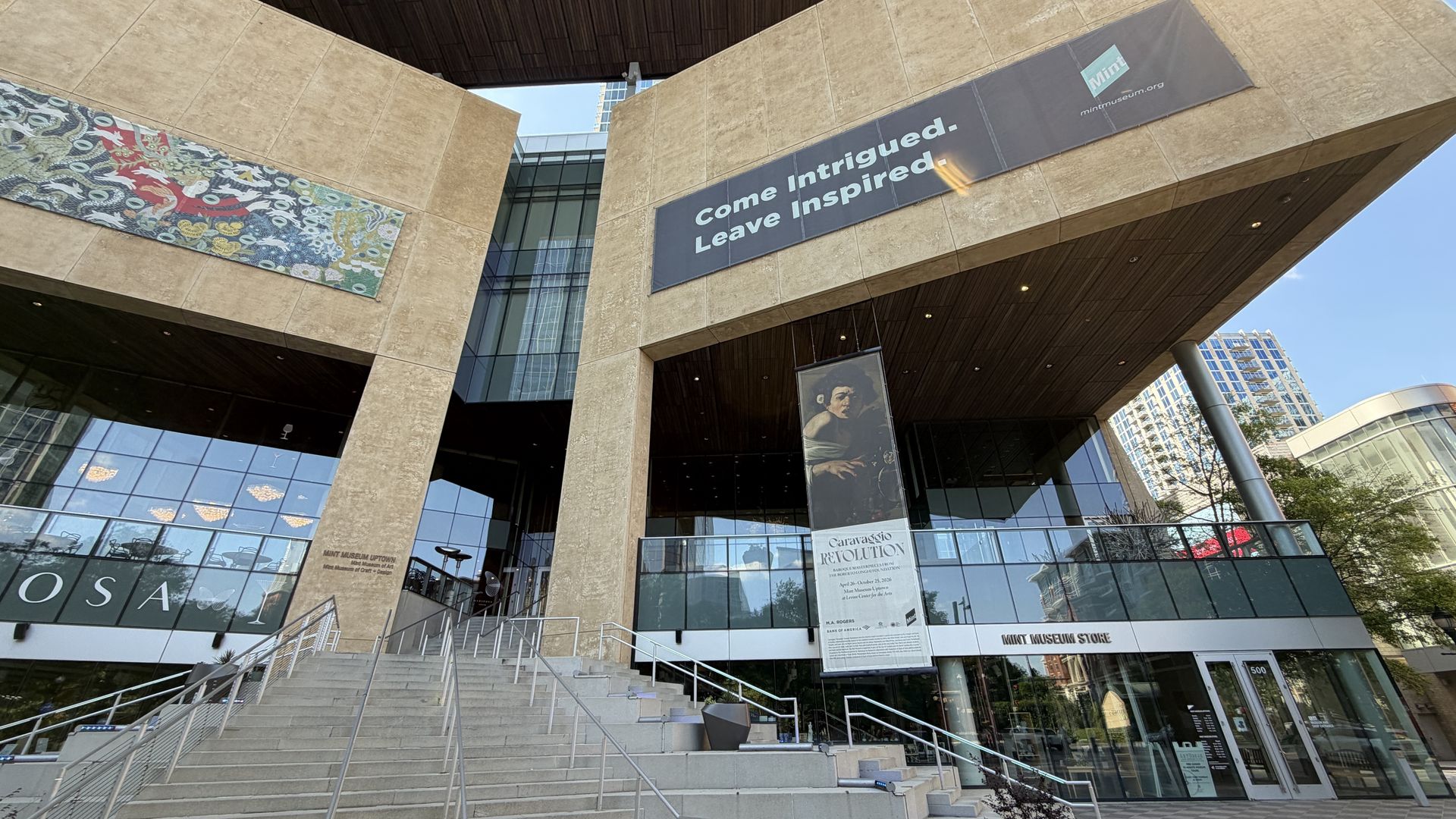 Exterior view of a modern museum entrance with beige stone walls, a banner reading "Come Intrigued. Leave Inspired." above glass doors, a colorful mosaic to the left, and wide stairs up to the entrance.