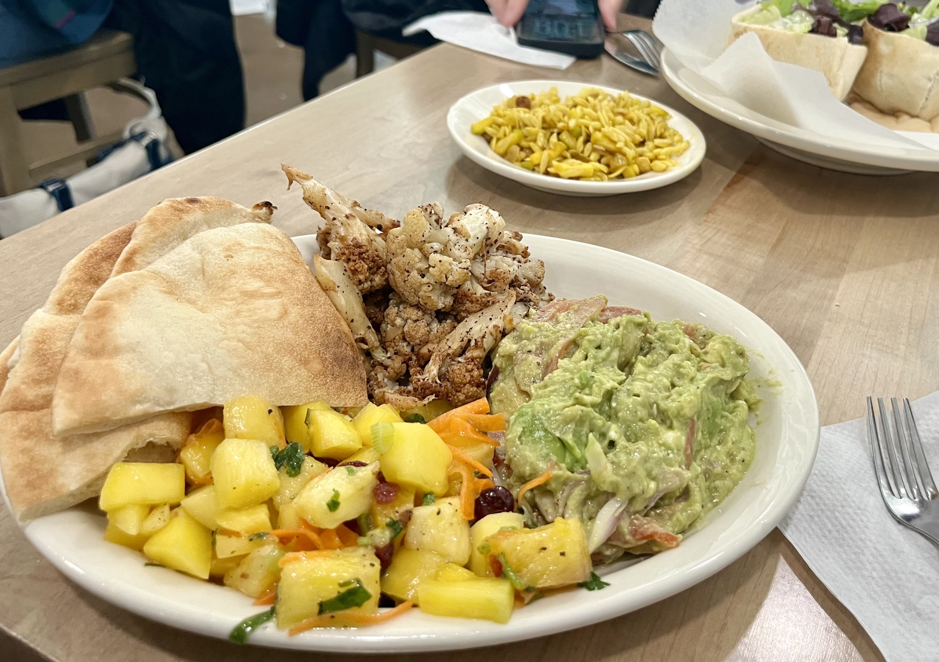 A large plate of food on a wooden table: roasted cauliflower with herbs, chunky avocado guacamole, mango-pineapple salsa, and warm flatbread; a yellow corn dish and other plates in the background.
