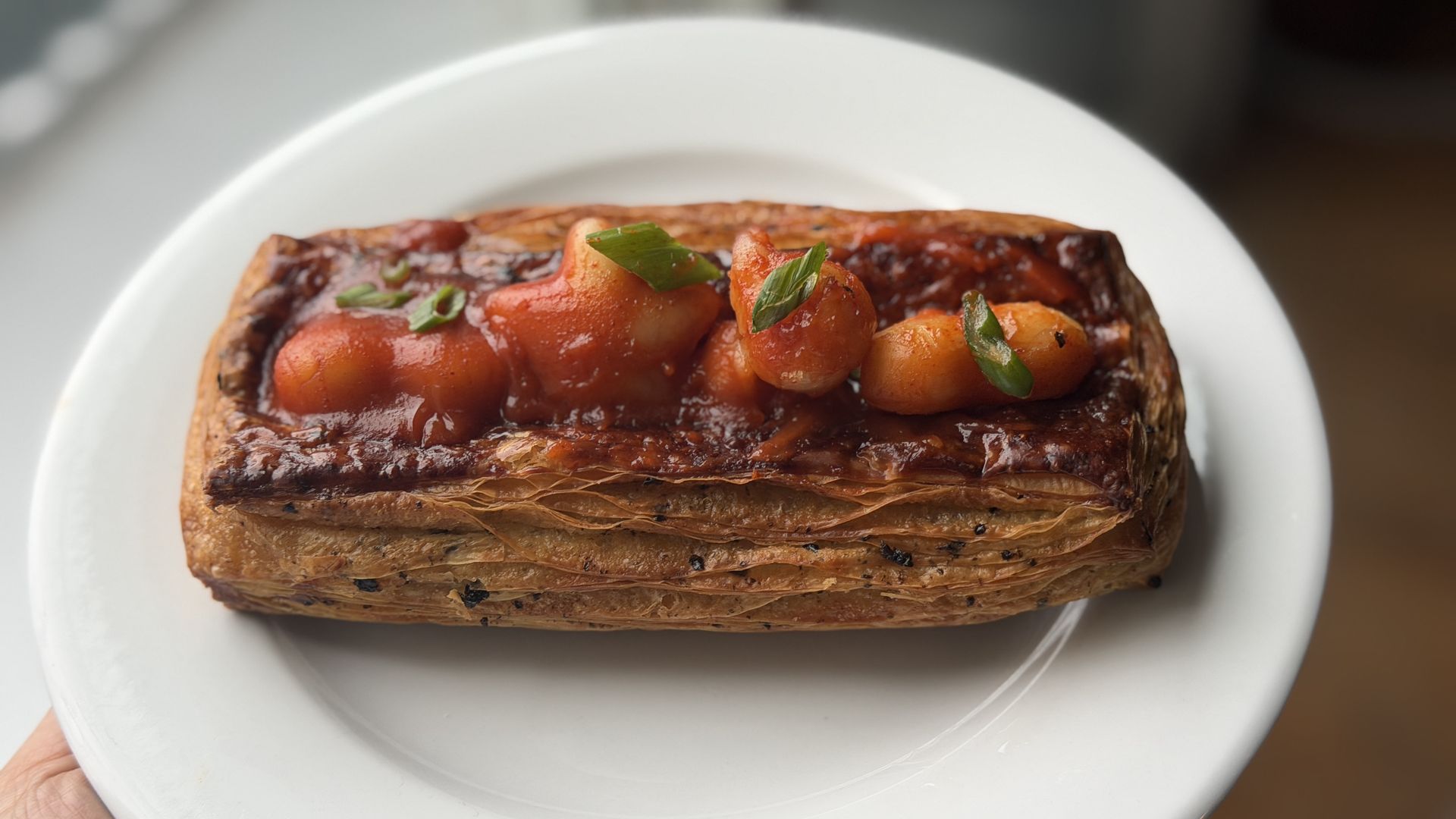 Rectangular puff pastry topped with baked beans in tomato sauce and chopped green garnish on a white plate, held by a hand near a window.