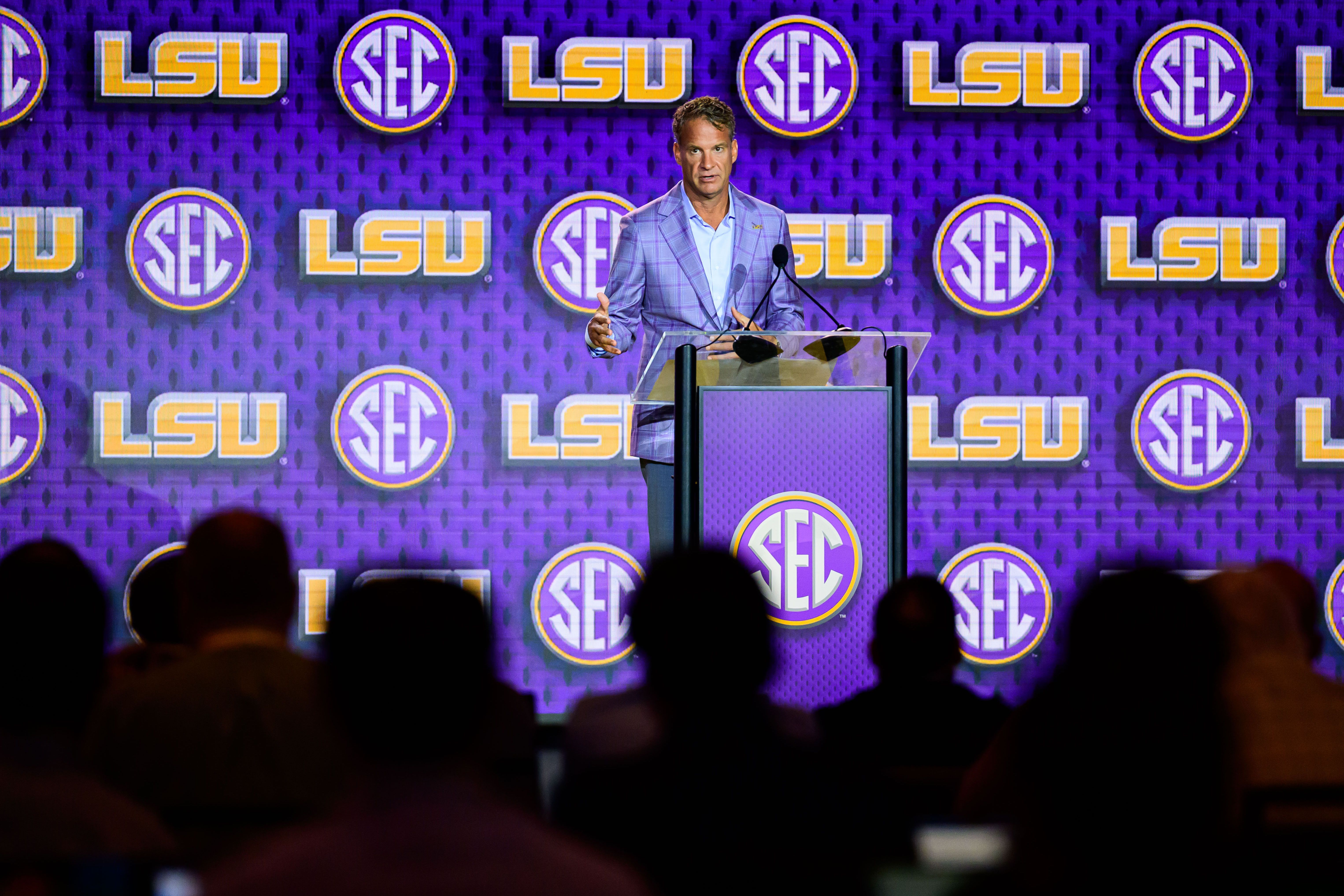 Speaker in a lavender plaid suit stands at a clear podium on a purple stage, with LSU and SEC logos in the background; silhouettes of the audience appear in the foreground.
