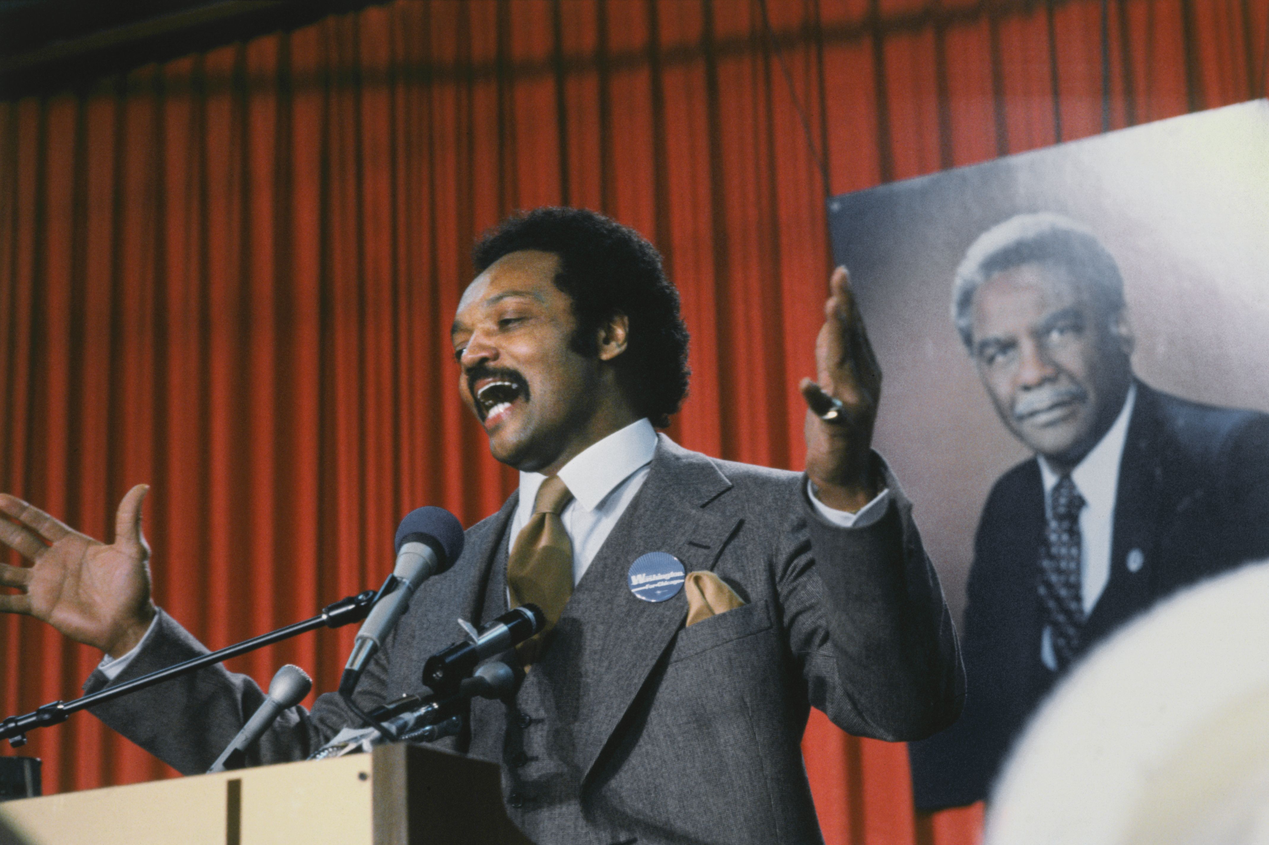 Man in gray suit and brown tie speaking energetically at a podium with microphones, gesturing with hands, red curtain background, portrait of older man in suit behind him.