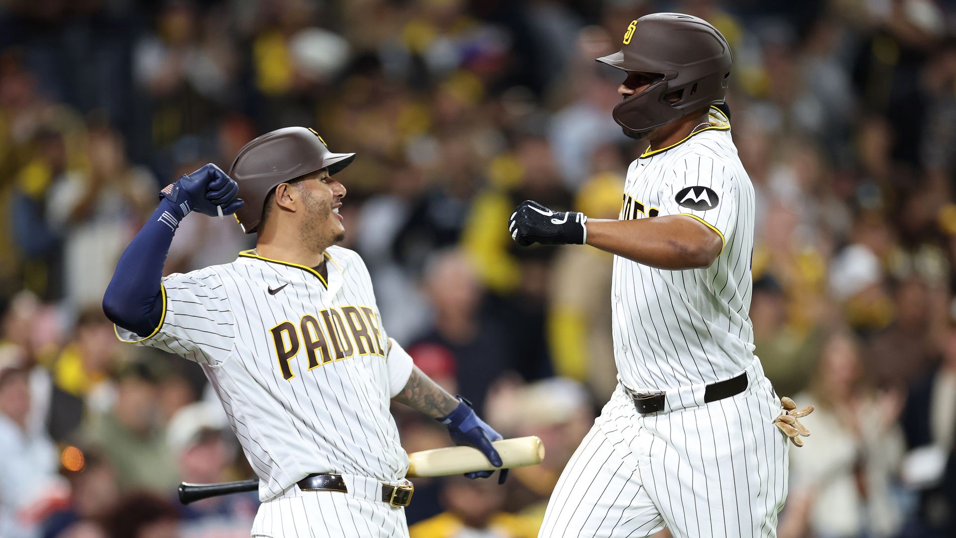 Two San Diego Padres players in white pinstriped jerseys celebrate on the field; left player raises a blue glove, right teammate leaps toward him for a high-five as a blurred crowd watches.