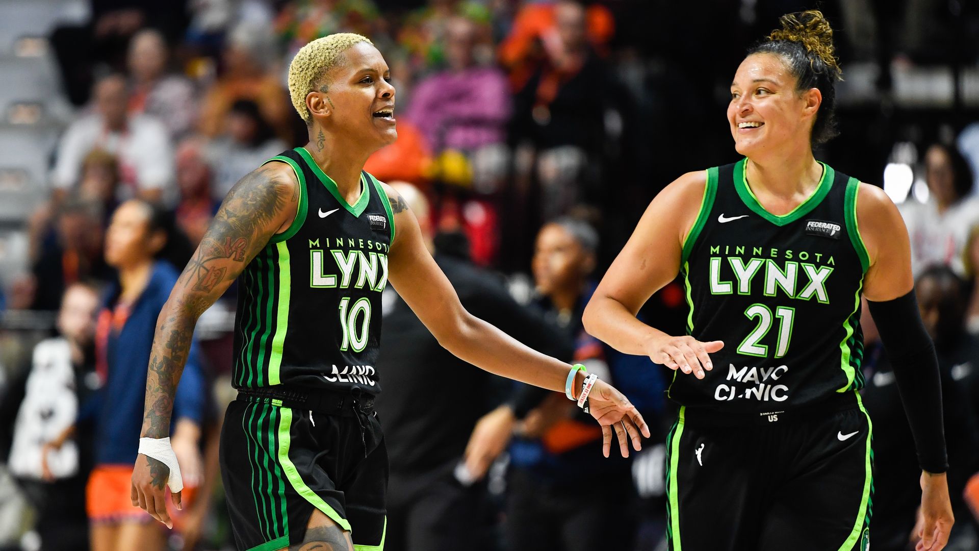 Two Minnesota Lynx players Courtney Williams and Kayla McBride in black jerseys with neon green trim smile as they jog on the court, with a blurred crowd in the stands behind them.