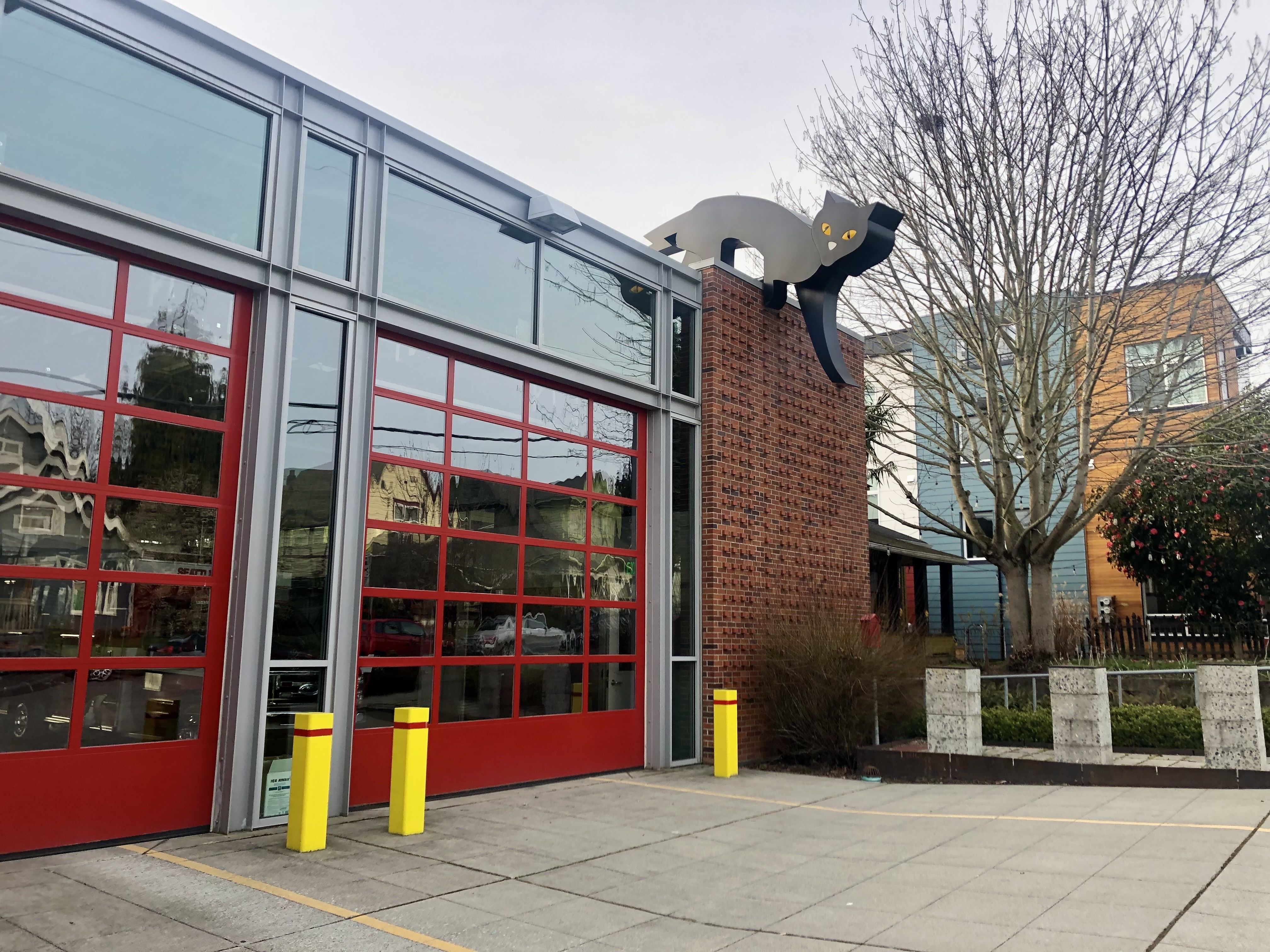 Modern building with tall glass panels and red-framed doors, brick wall, and a metal cat sculpture perched atop the wall with yellow eyes; leafless trees and cloudy sky behind.