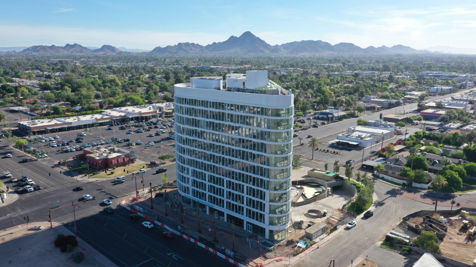 A modern curved glass office building under construction in a sunny urban area with mountains in the background and surrounding roads, parking lots, and greenery.
