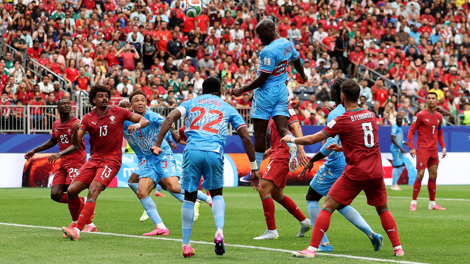 Soccer match: players in red and light-blue jerseys jump for a ball amid a packed stadium, with a sea of red-clad fans in the stands.