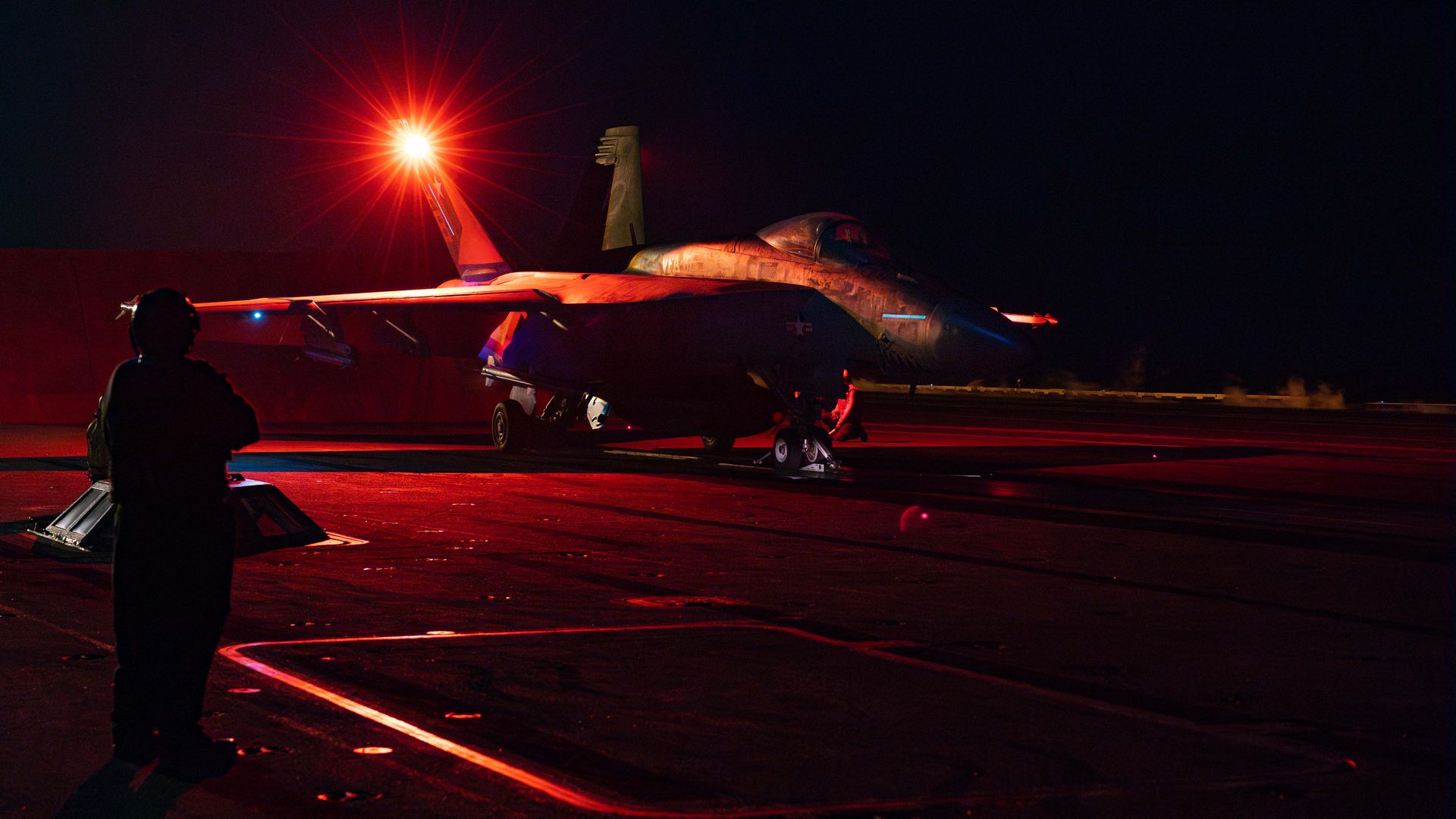 An American fighter jet sits on the deck of an aircraft carrier. A person stands to the left of the foreground. It's night time, and everything is bathed in red light.